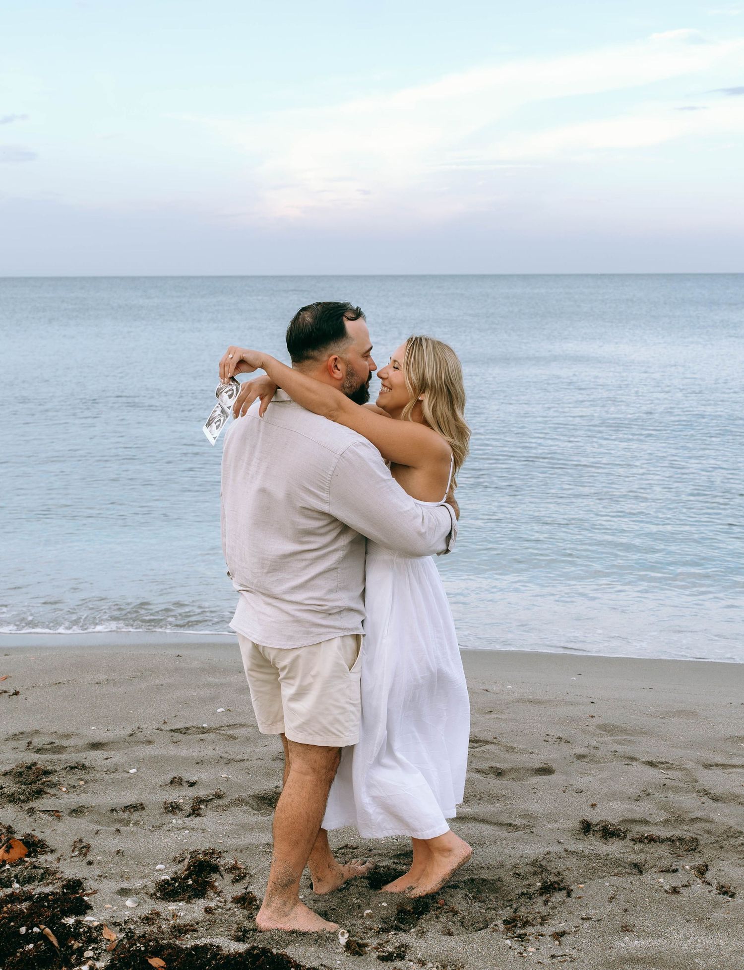 Couple embracing and kissing on beach at sunset in coordinating white and beige outfits.