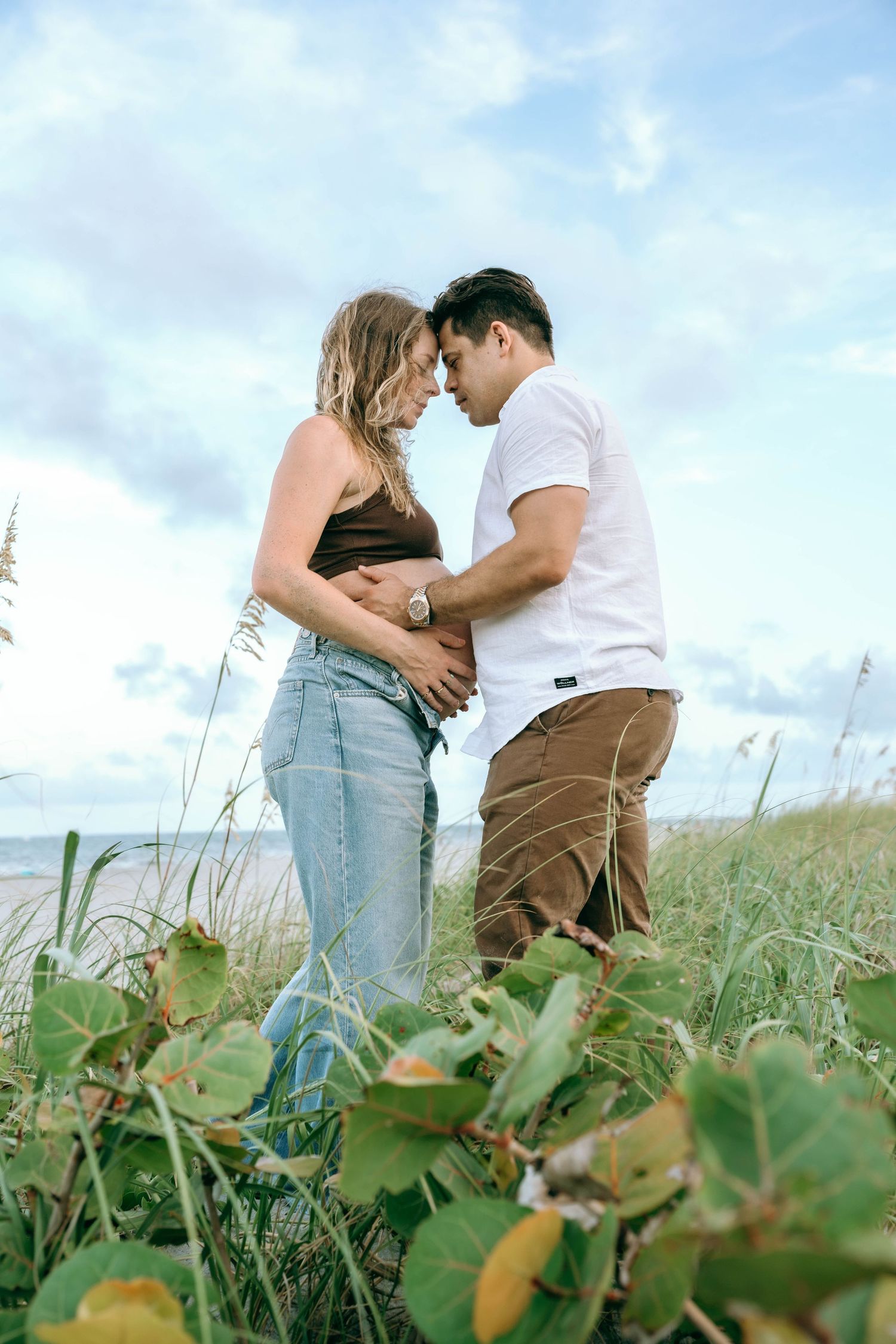 A couple shares a tender forehead touch outdoors, the woman visibly pregnant, both cradling her belly near a coastal grassy area.