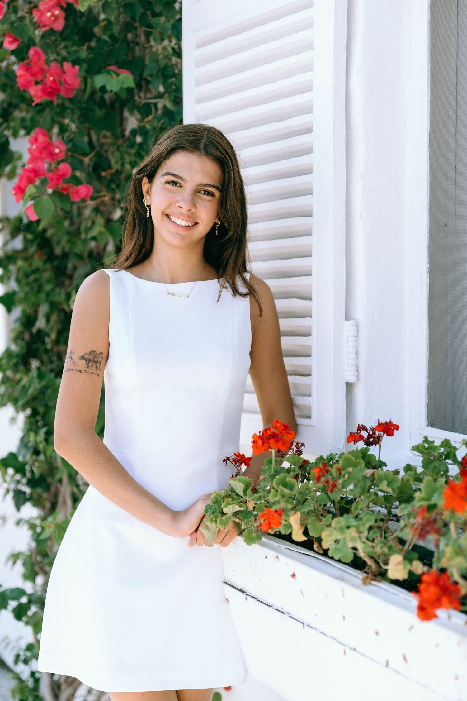 Young woman in white mini dress smiling beside red flowers and white shuttered window on Worth Avenue Palm beach.