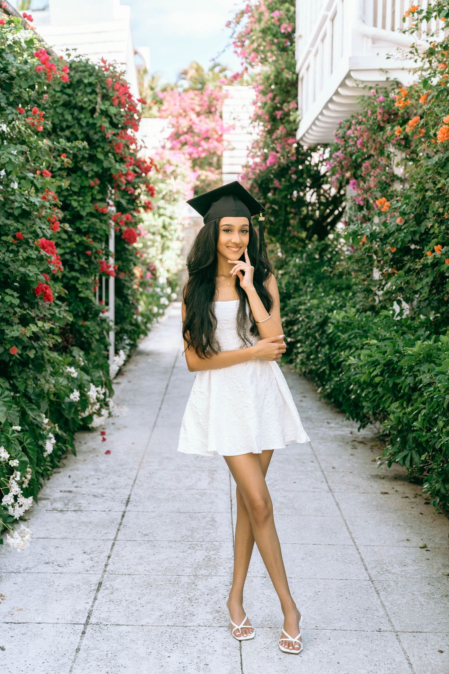 Young woman in white dress and graduation cap posing in a flower-lined pathway on Worth Avenue Palm beach.