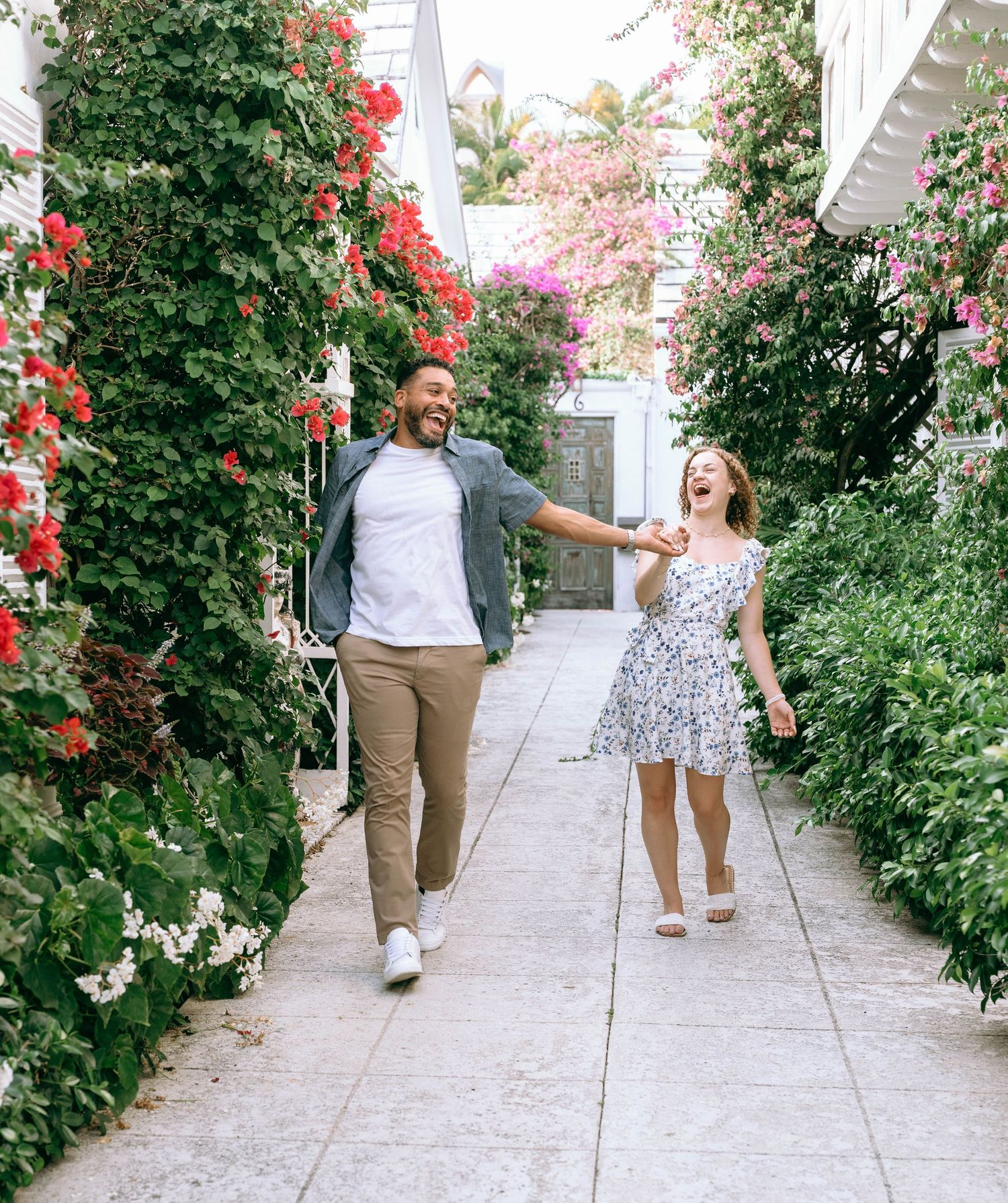 A joyful couple holds hands while walking through a flower-lined pathway with red and pink blooms. Engagement photos on Worth Avenue in Palm