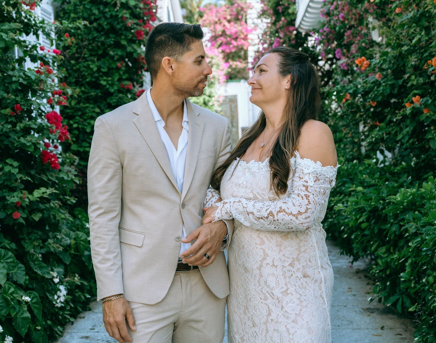 Couple in wedding attire gazing at each other in a floral garden pathway, groom in beige suit, bride in white lace gown.