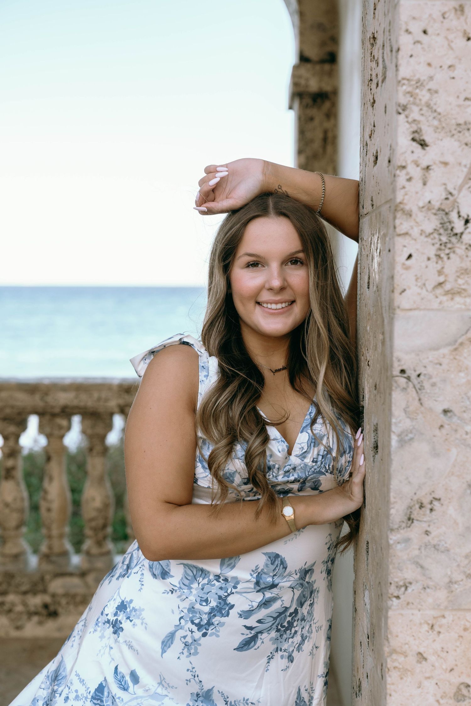 Smiling woman in blue floral dress leans against stone column with ocean view behind her.