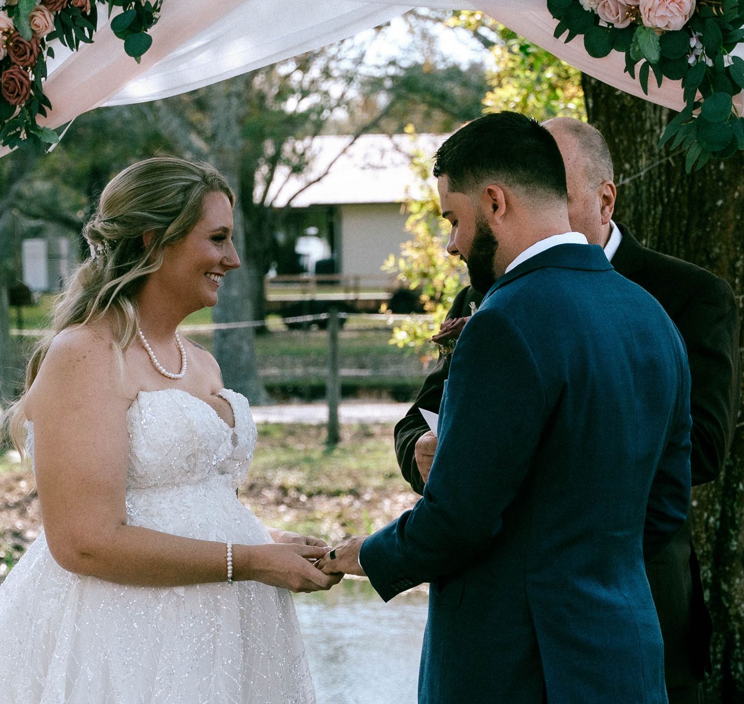 Bride and groom exchanging vows outdoors under floral arch, bride in lace gown smiling at groom in navy suit.
