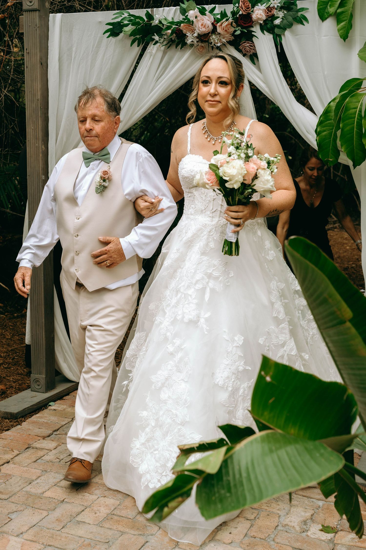 A wedding party walks along a brick path lined with tropical foliage and white drapery.