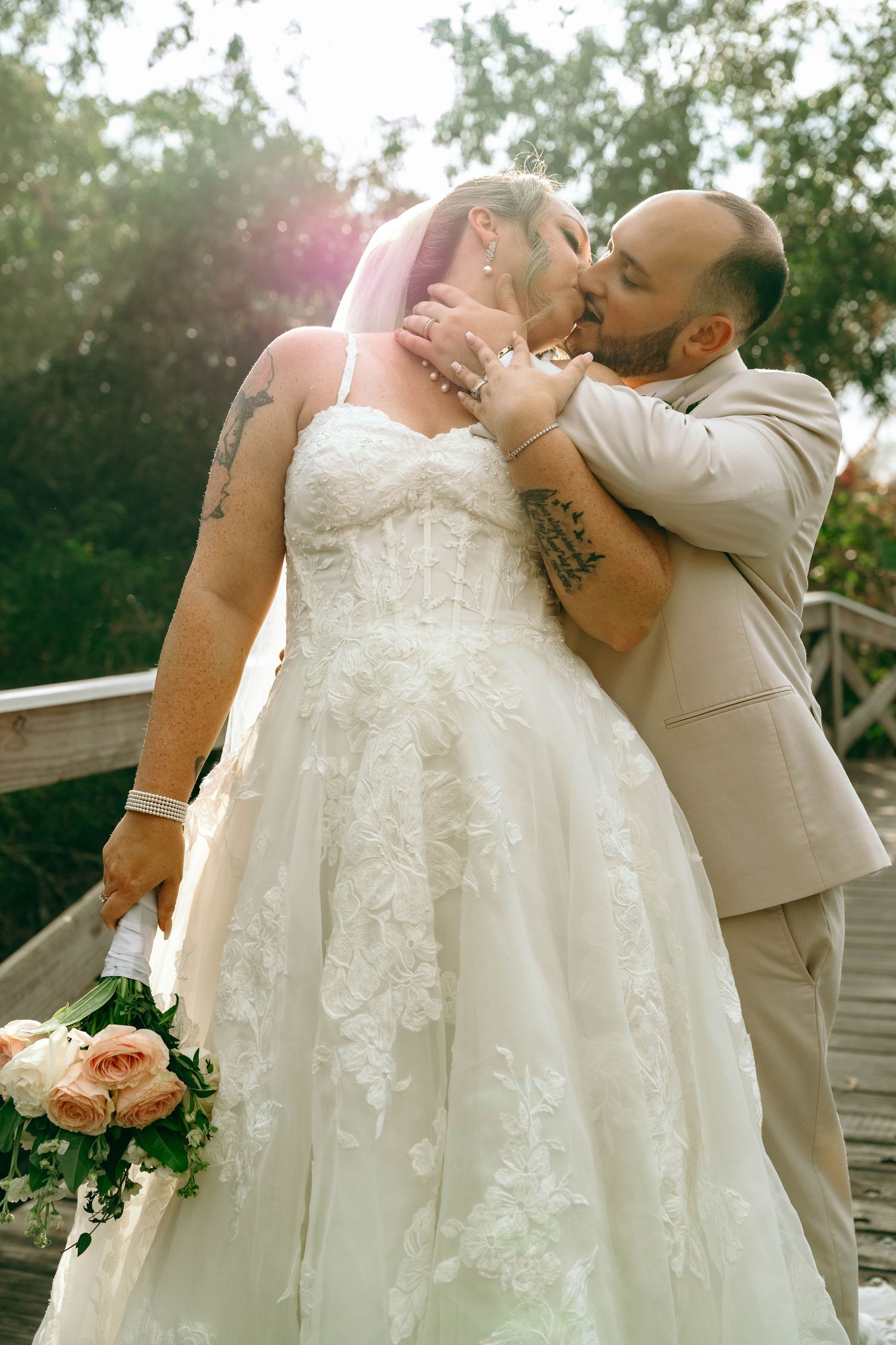 A romantic wedding moment captured outdoors with a bride in a lace gown and groom in beige suit sharing a tender kiss.