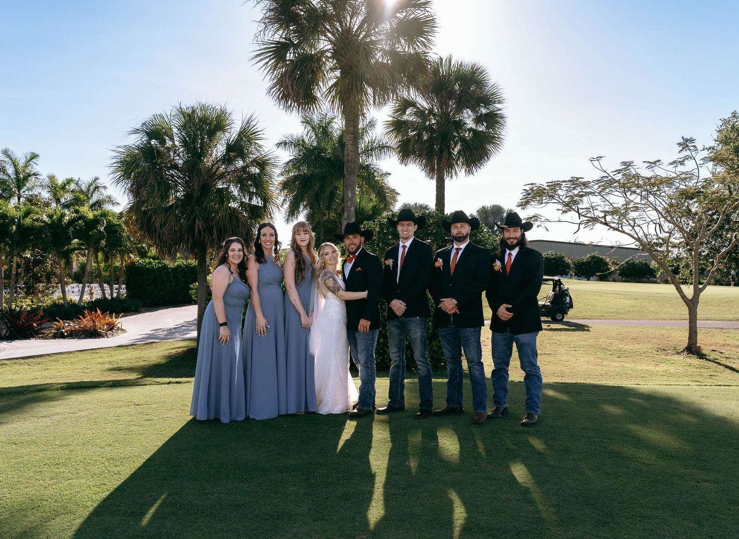 Wedding party poses together on sunny lawn with palm trees in background.