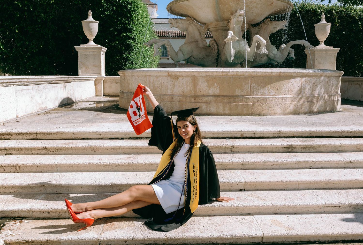Graduate in cap and gown holding red flag while sitting on fountain steps at outdoor ceremony on Worth Avenue Palm beach.