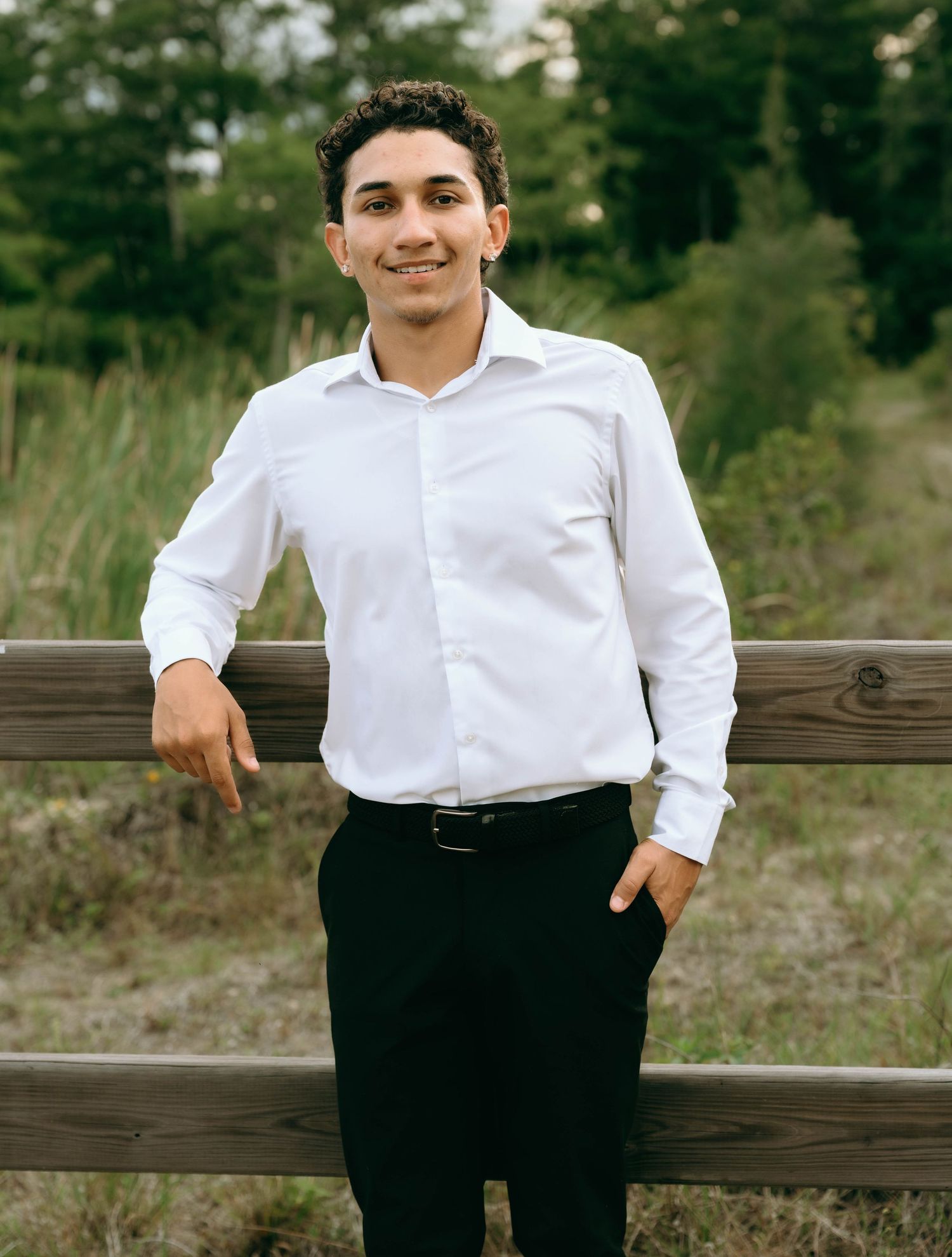 Smiling young man in white dress shirt and black pants leans against wooden fence in nature setting. Senior Photos at Colt Farms in Boynton