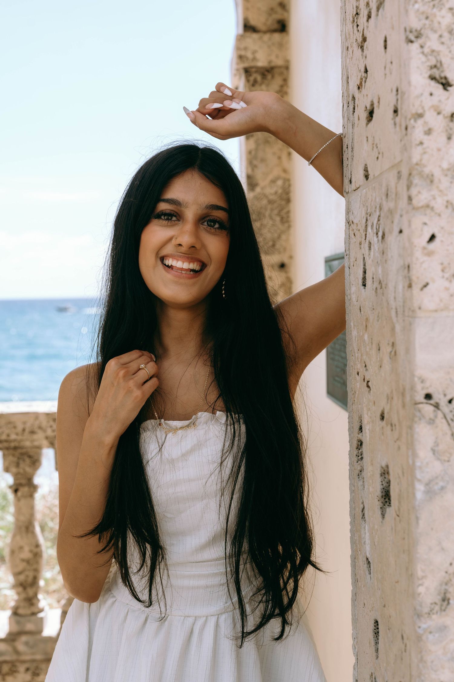 Smiling young woman with long black hair in white dress posing by stone column with ocean view on Worth Avenue Palm beach.