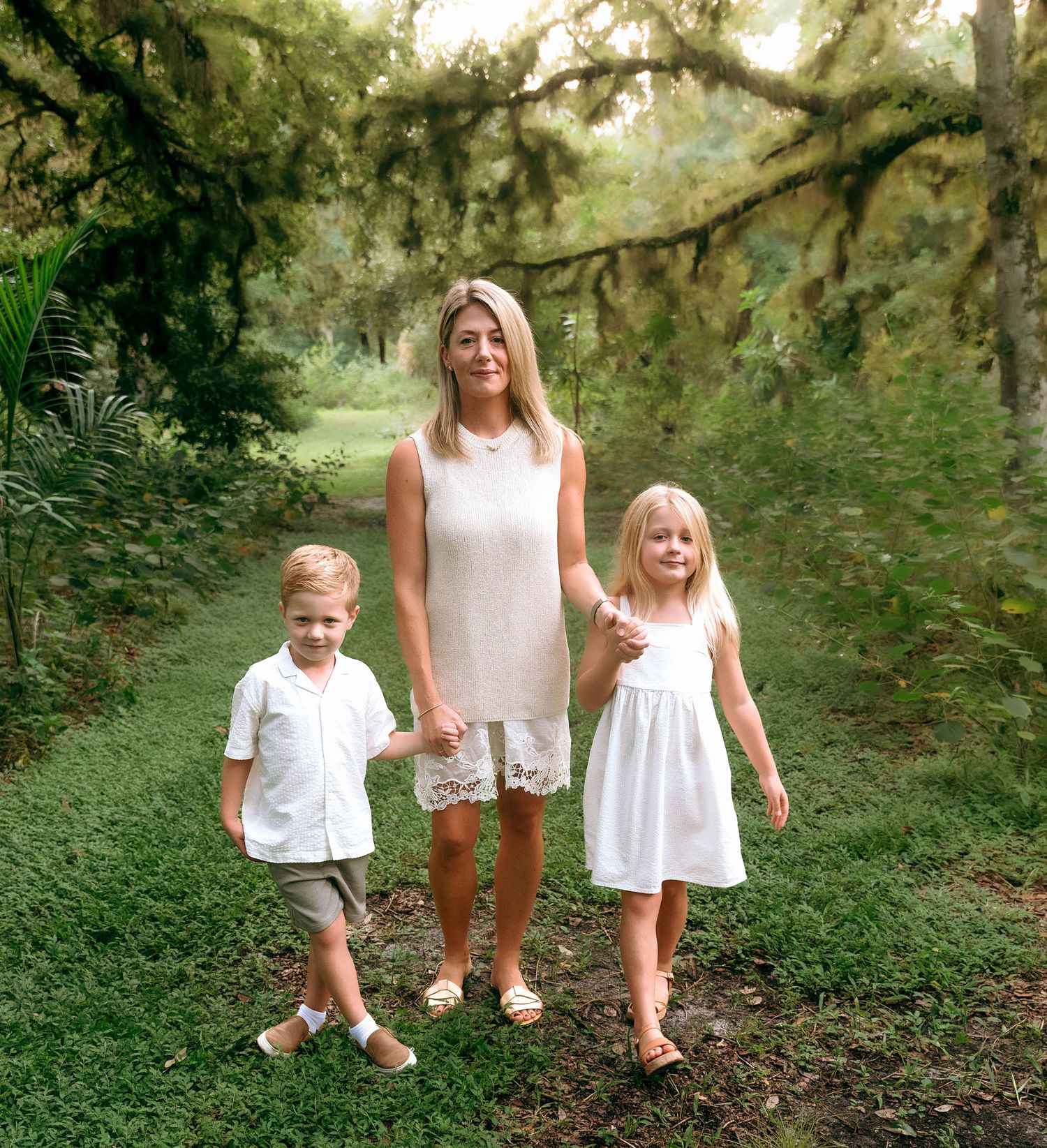 A woman in a white lace dress holds hands with two young children in white outfits on a lush green garden path at Riverbend Park in Jupiter,