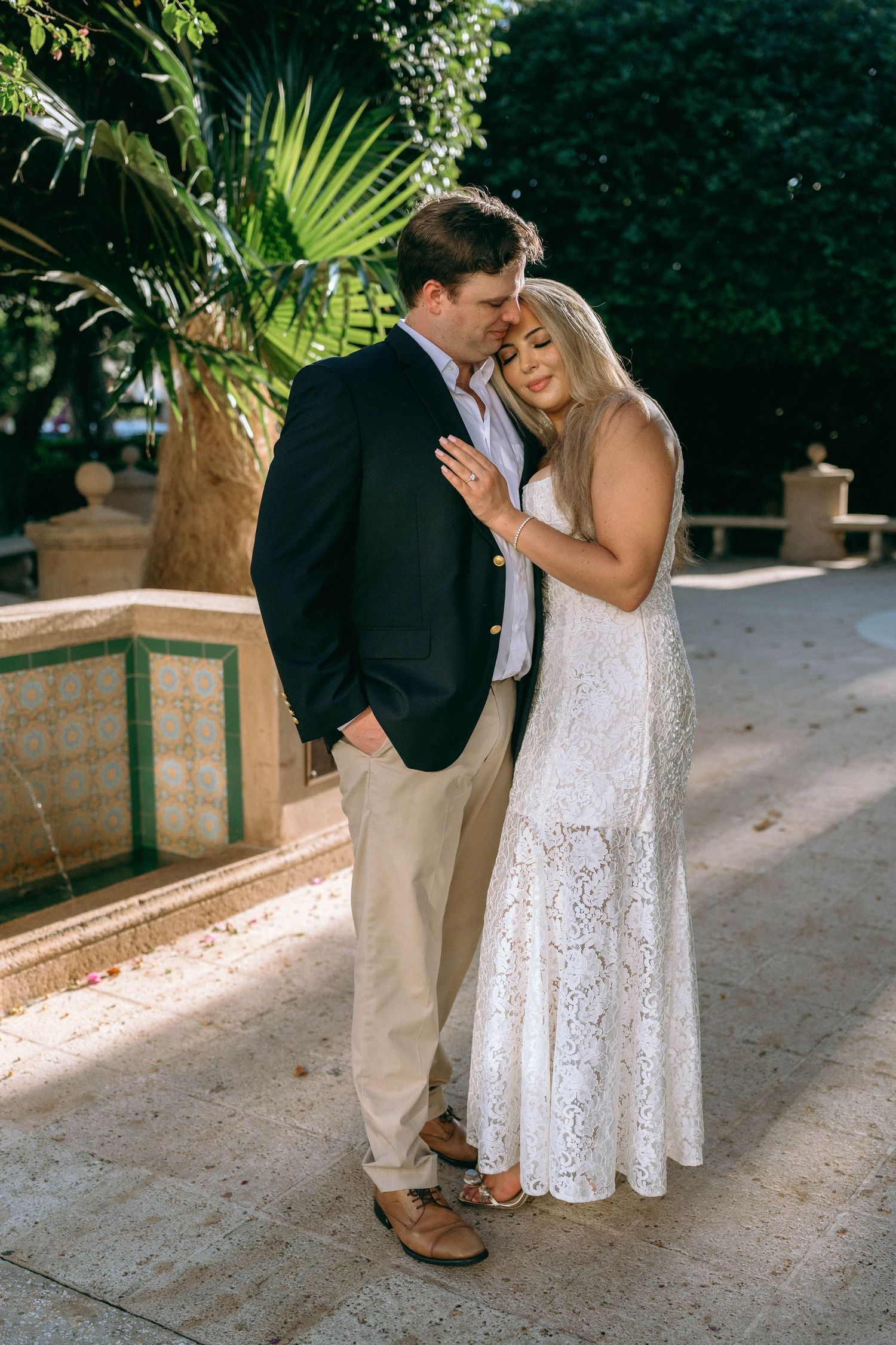 Couple embracing outdoors in a garden, woman in white lace dress, man in navy blazer and khakis, tropical plants background. Worth Avenue