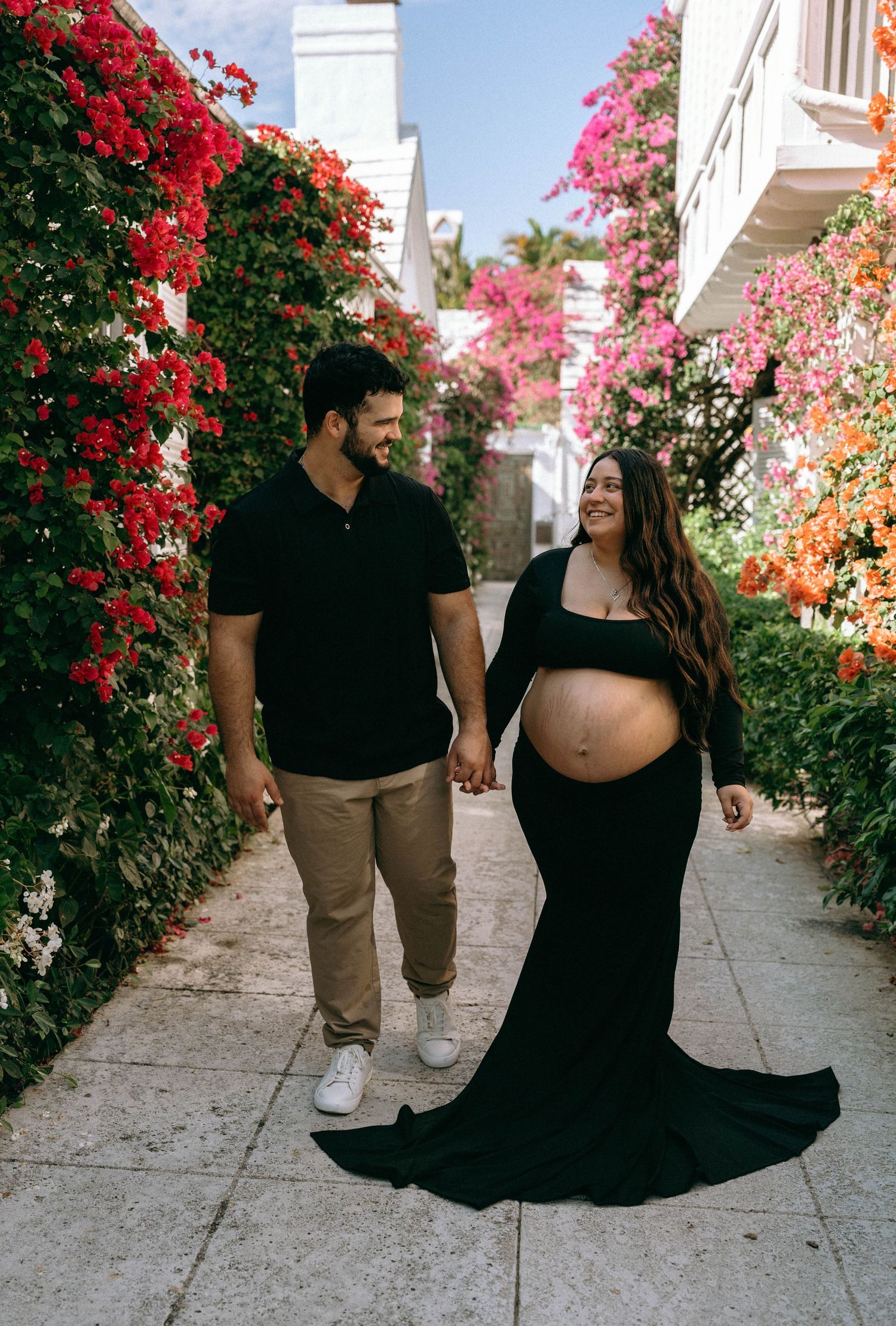 Happy expecting couple walks hand-in-hand through flower-lined pathway with pink and red blooms. Worth Avenue, Palm Beach, FL