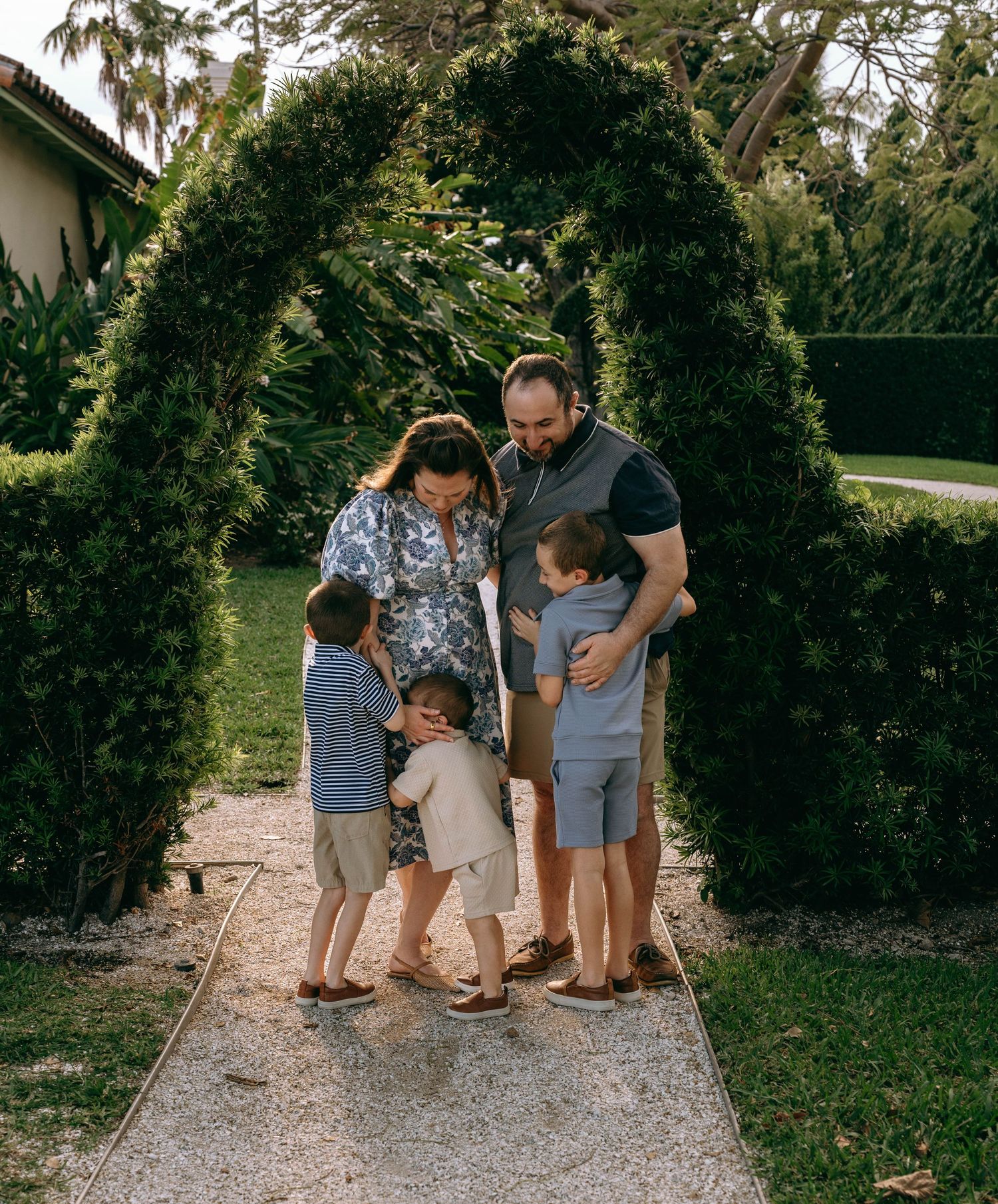 Family of five sharing a warm embrace under a garden arch, children playfully hugging parents on a stone path.  Family photos Bradley Park