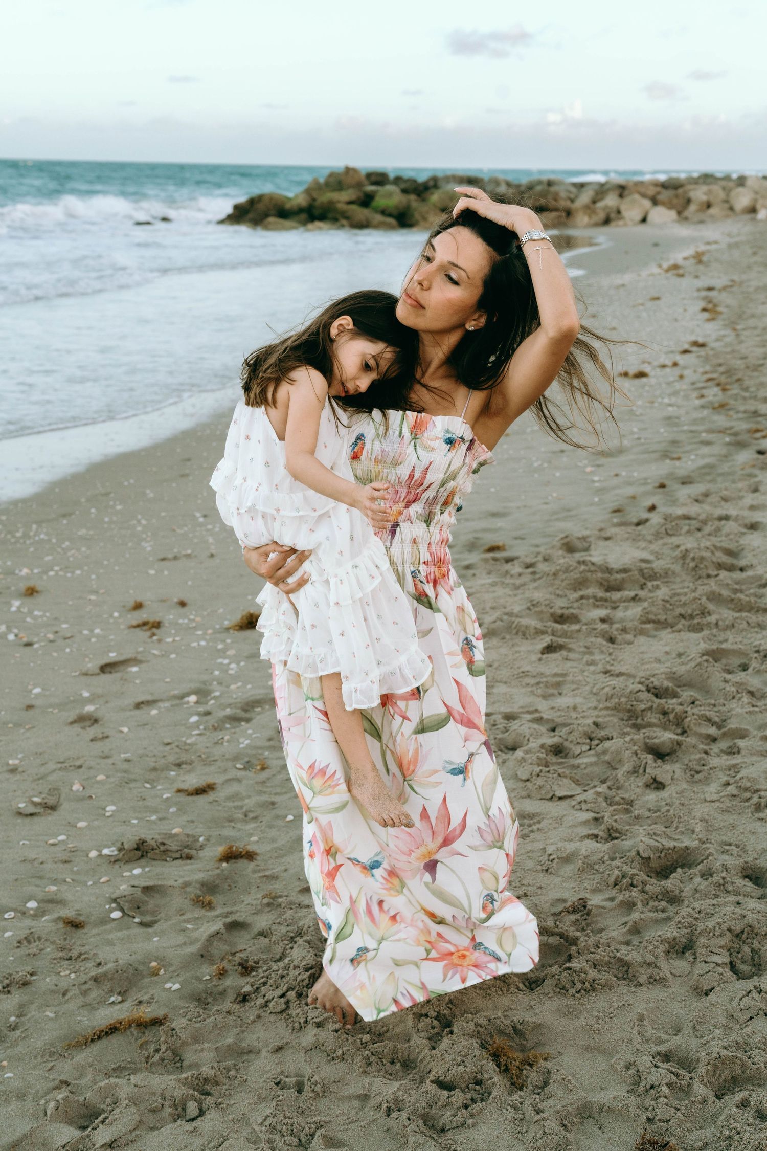 Mother in floral maxi dress holds young daughter on sandy beach, both wearing matching white floral outfits. Worth Avenue, Palm Beach, FL