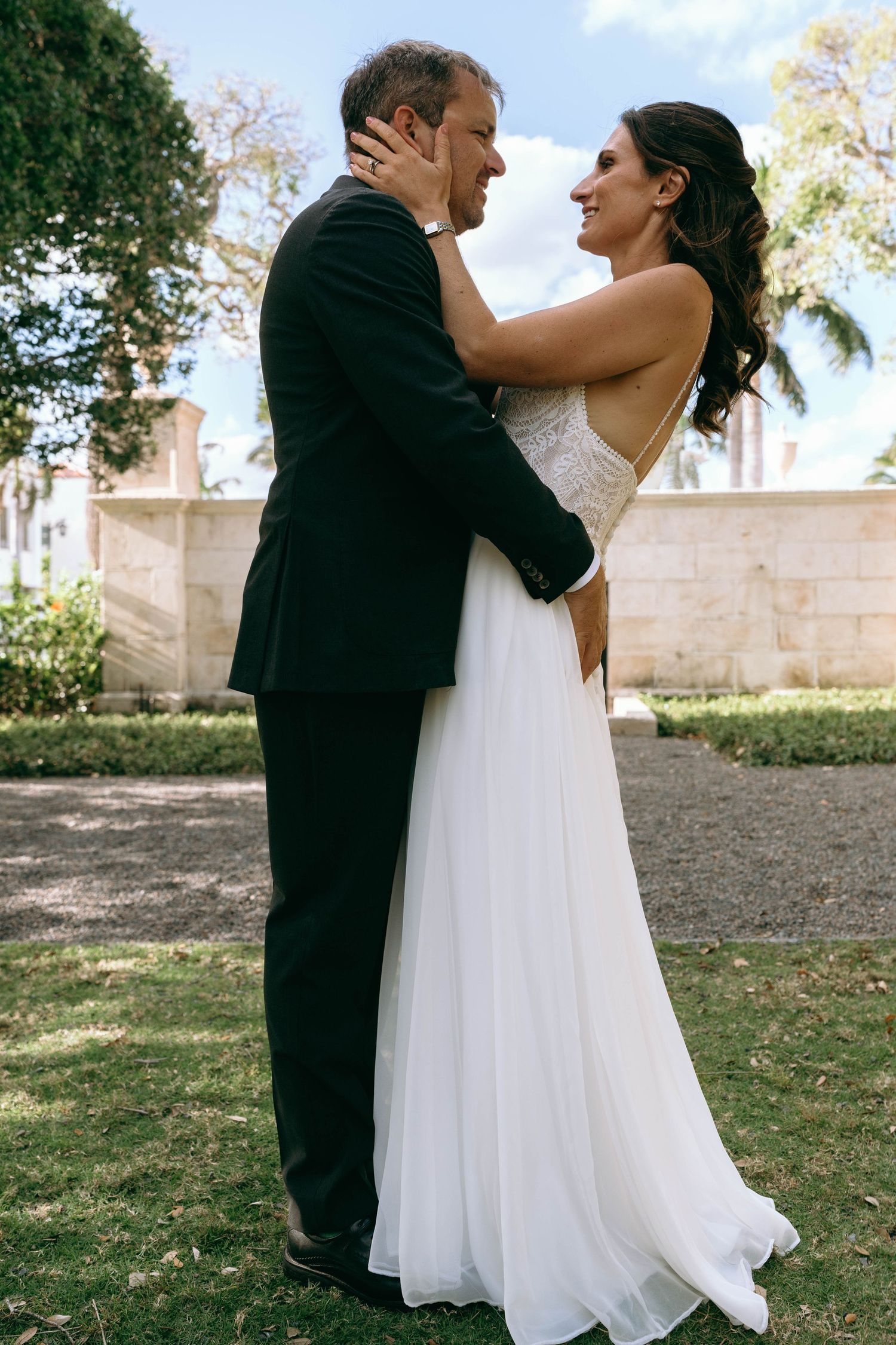 Bride and groom share a romantic embrace outdoors, with the bride in a white backless gown holding the grooms face.