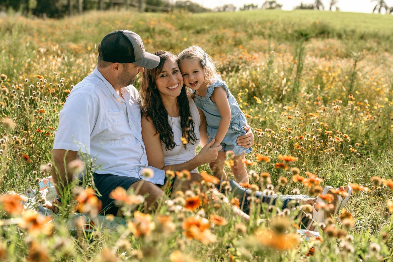 Family of three smiling together in a sunny wildflower field, parents holding young daughter among orange blooms.