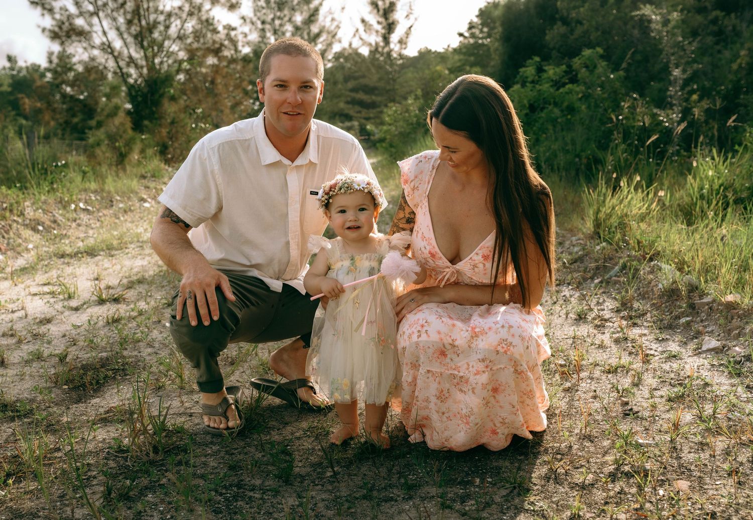 Family of three poses outdoors, parents smiling with toddler girl in floral dress and flower crown on sandy path.