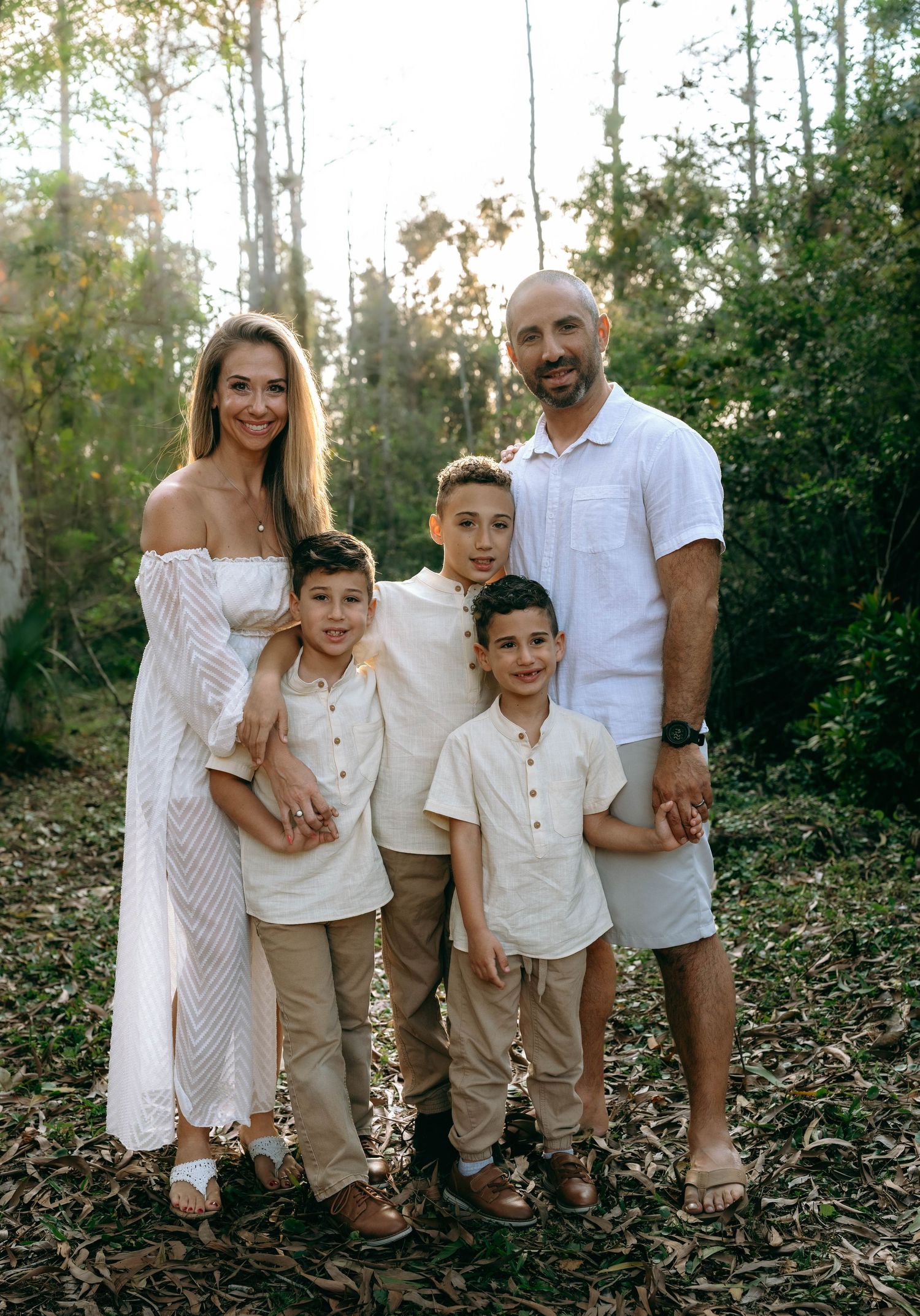 Family wearing coordinated white and beige outfits poses together in a natural outdoor setting among trees and foliage.