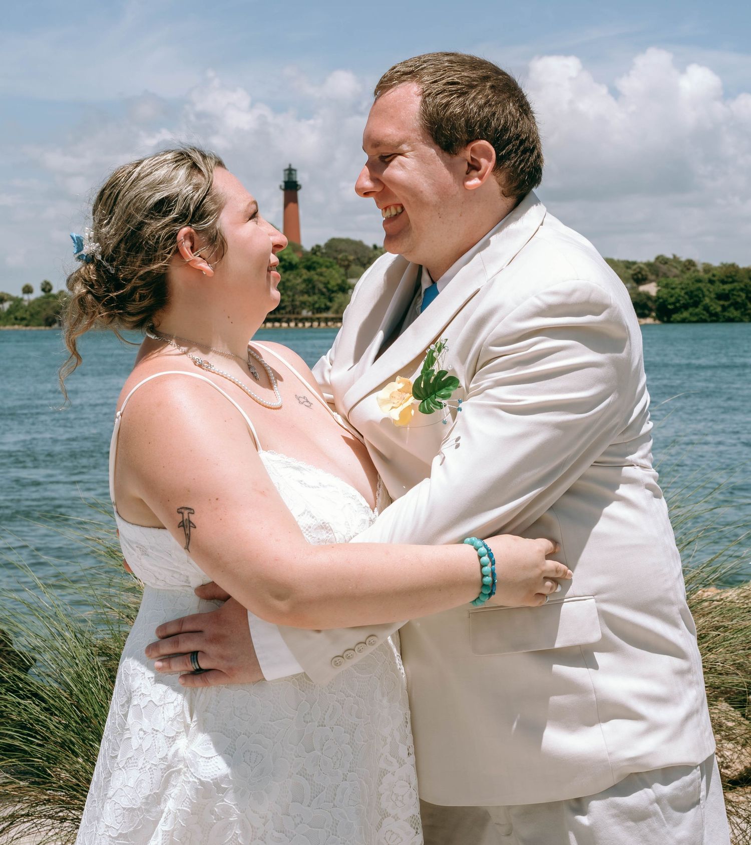 Smiling bride and groom embrace near waterfront lighthouse backdrop at outdoor summer wedding.