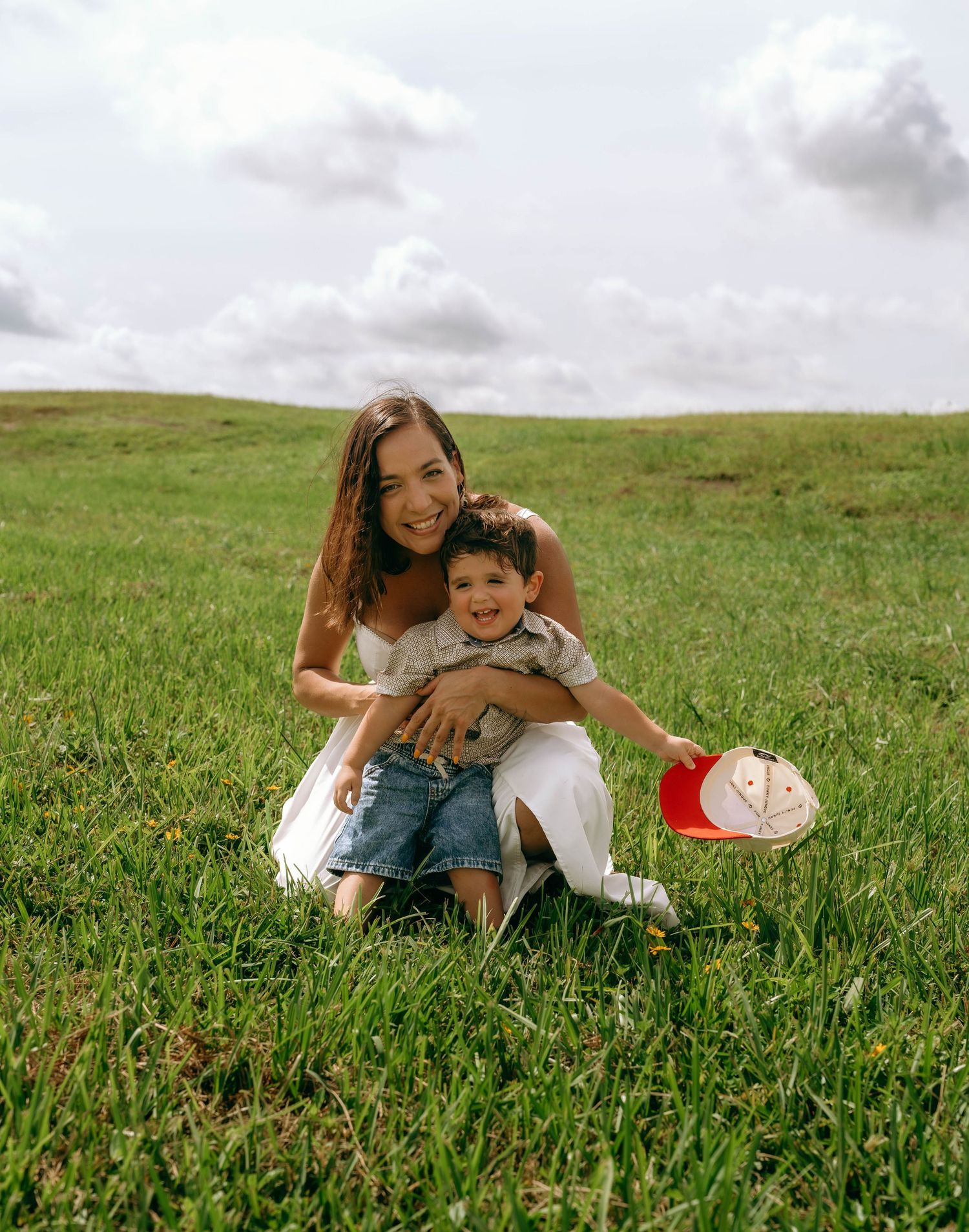 A smiling woman sits on green grass holding a young boy with a red cap on a cloudy day.