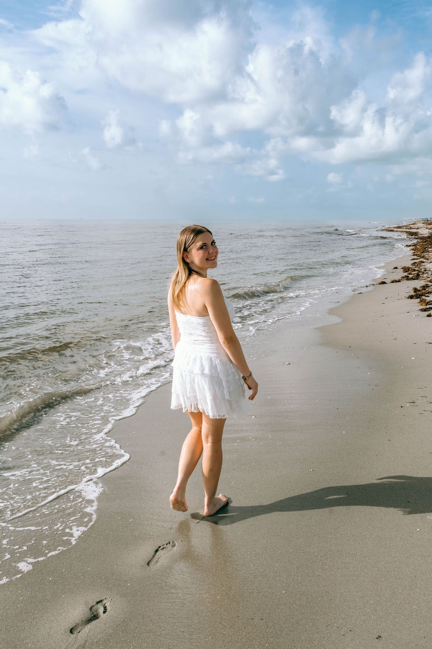 Young woman in white dress walking barefoot on sandy beach with ocean waves and cloudy blue sky.