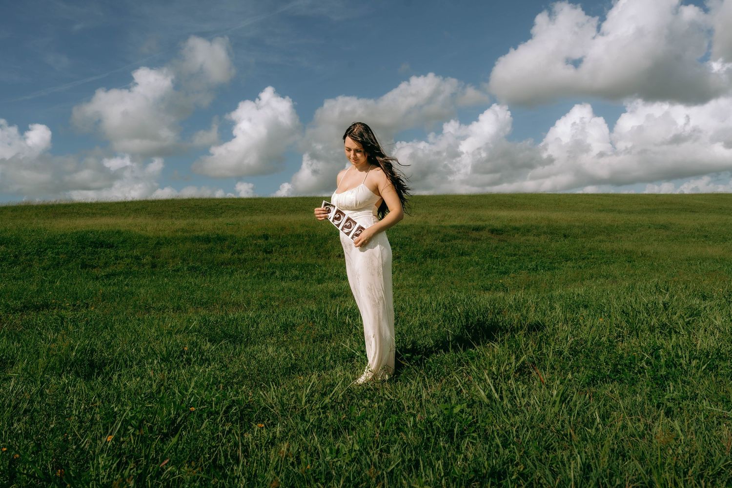 Pregnant woman in white dress holding baby shower sash in lush green field under blue sky with clouds.