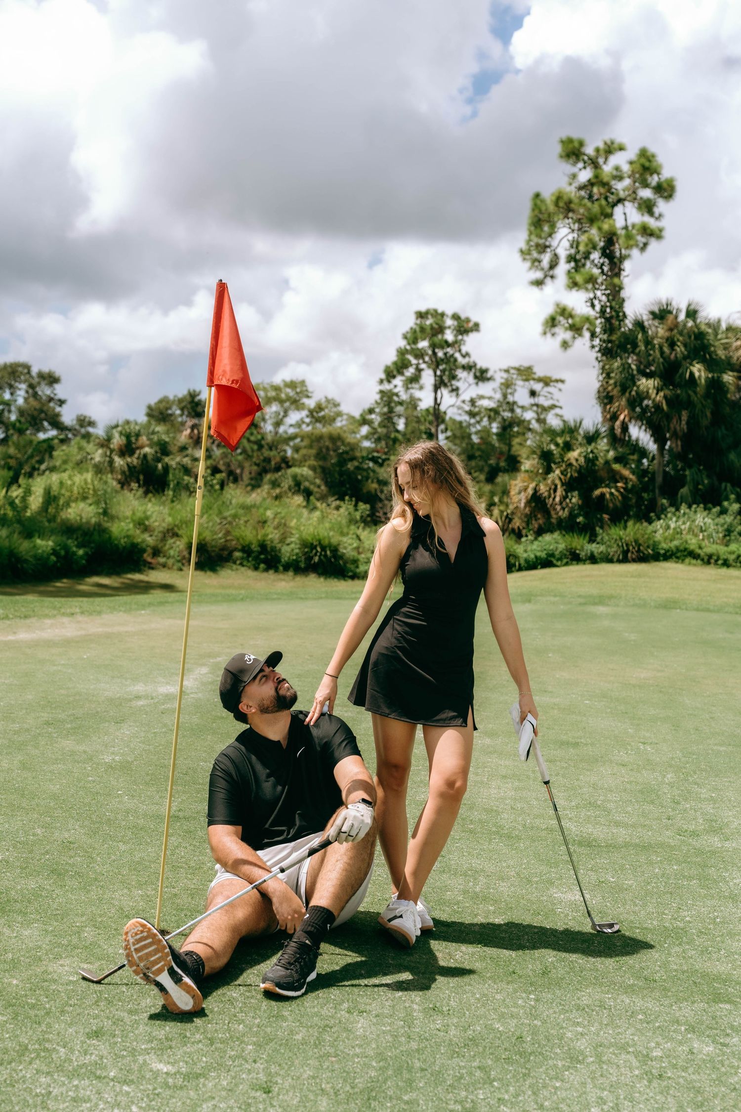 Couple on golf course green, woman in black dress holding club, man sitting by red flag, tropical trees background.