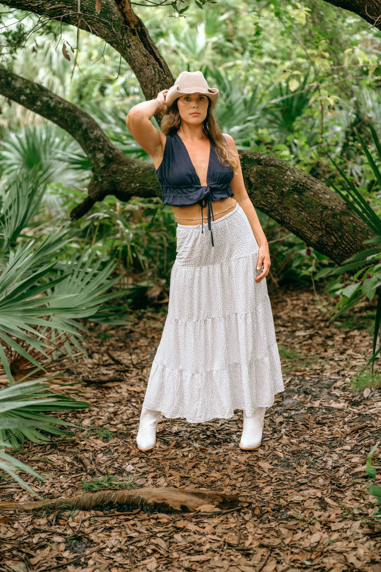 Woman in white maxi skirt, navy crop top, and tan hat poses in lush tropical forest setting.
