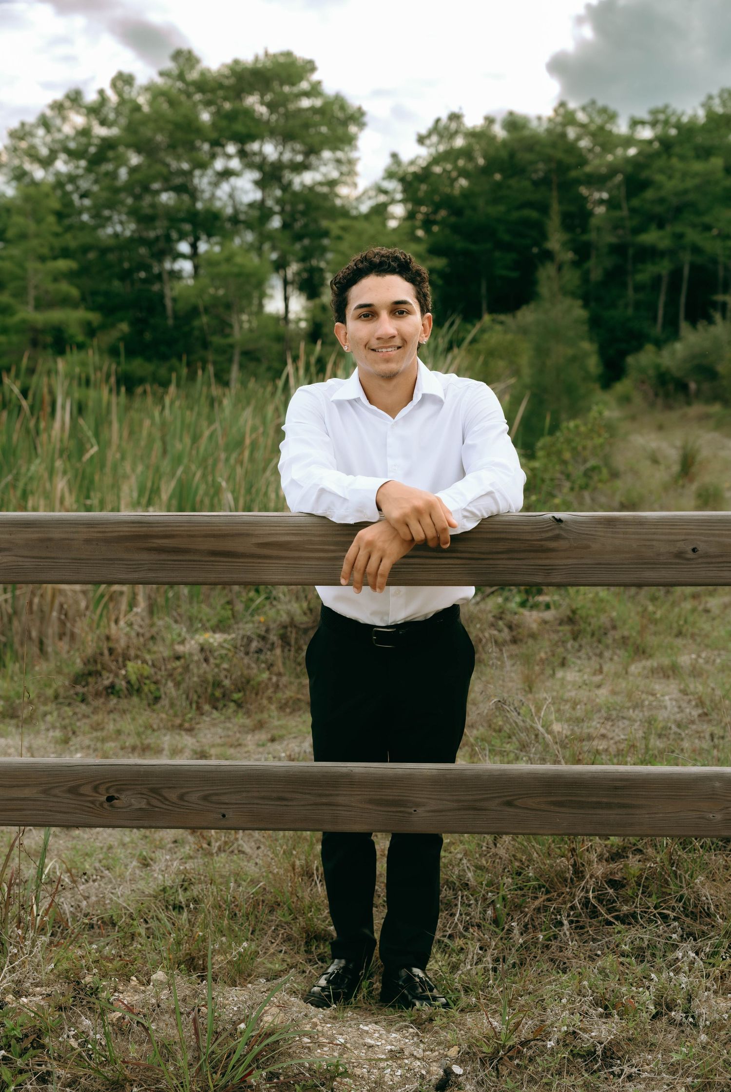 Smiling young man in white dress shirt leans on wooden fence rail in a natural outdoor setting. Senior Photos at Colt Farms in Boynton Beach