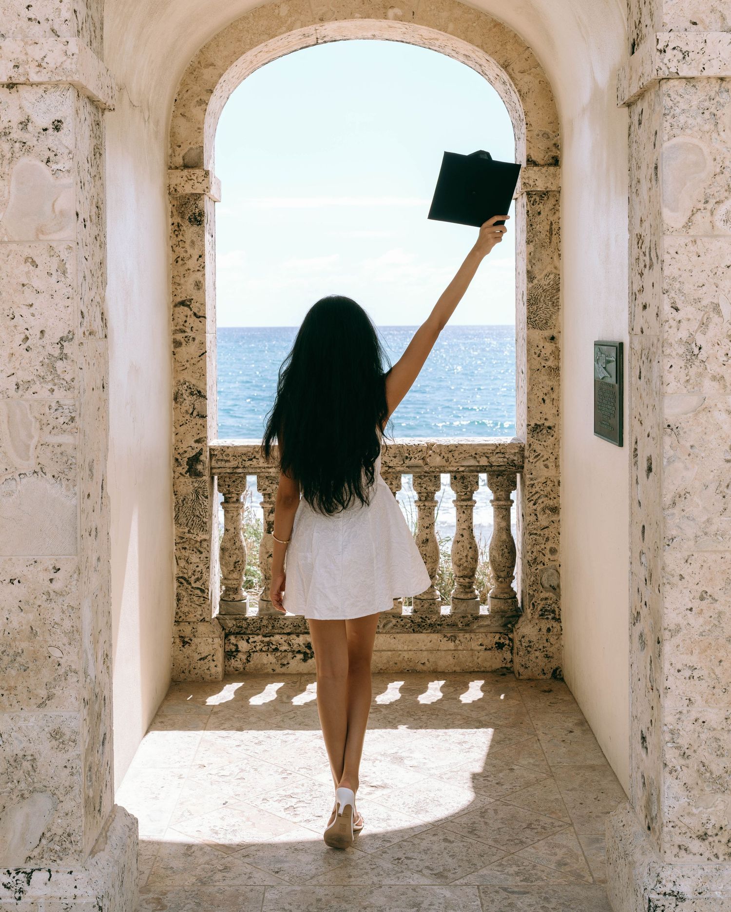 Young woman in white dress raising graduation cap under stone arch overlooking the ocean on Worth Avenue Palm beach.