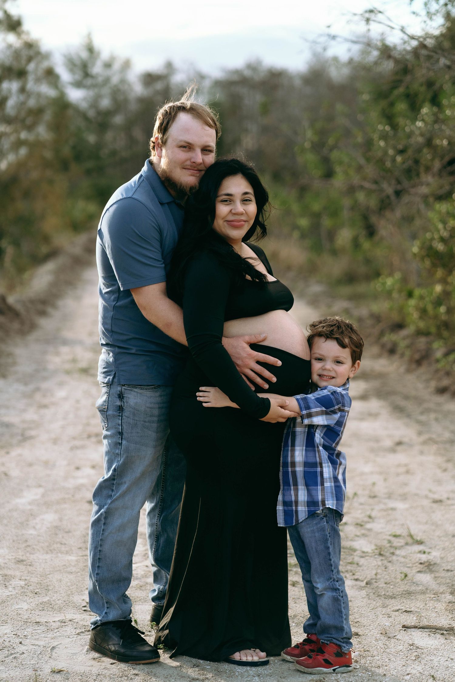 A maternity photo on a dirt path shows a family embracing a pregnant woman in a black dress.