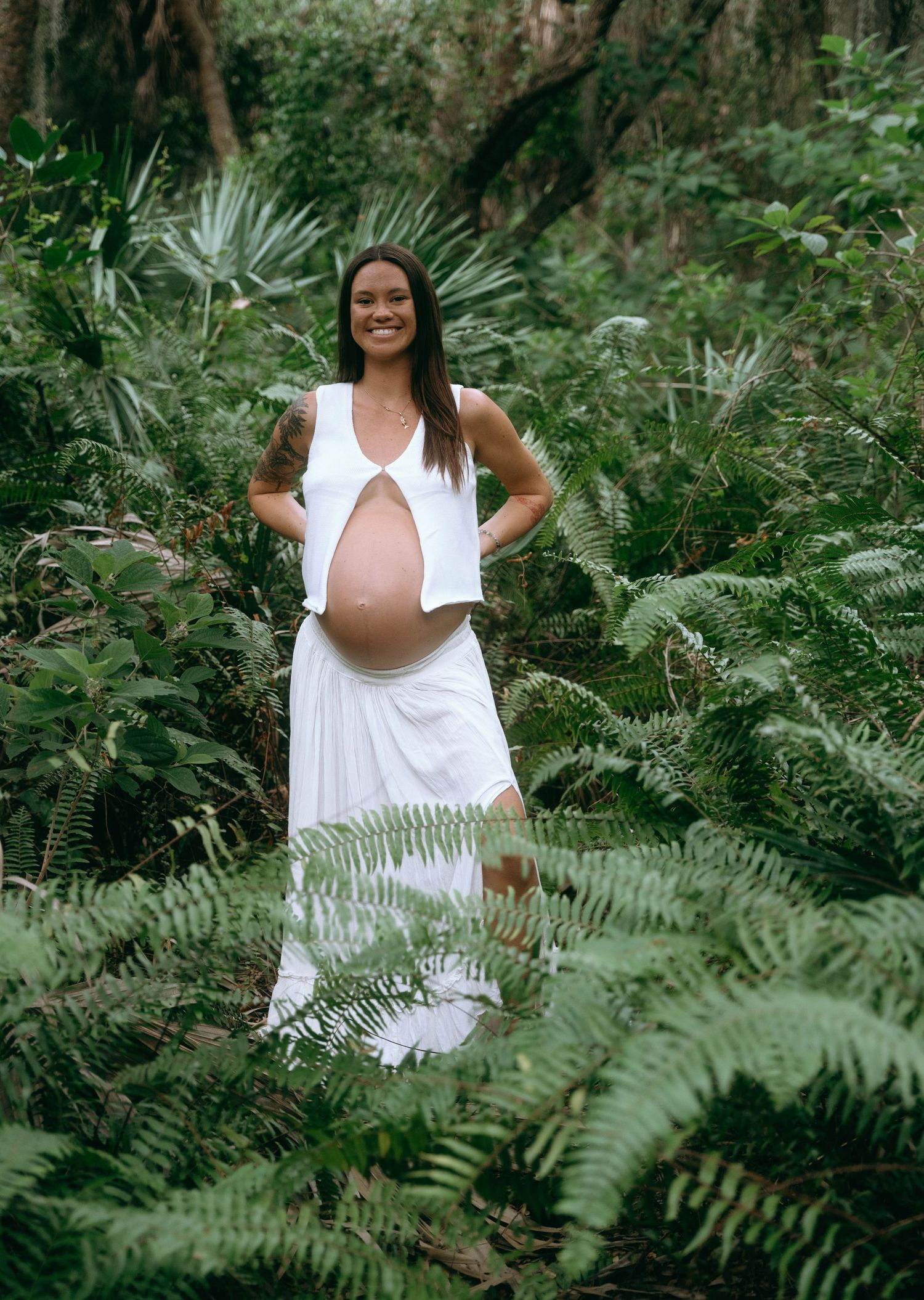 A maternity portrait in a lush fern garden wearing a flowing white dress.