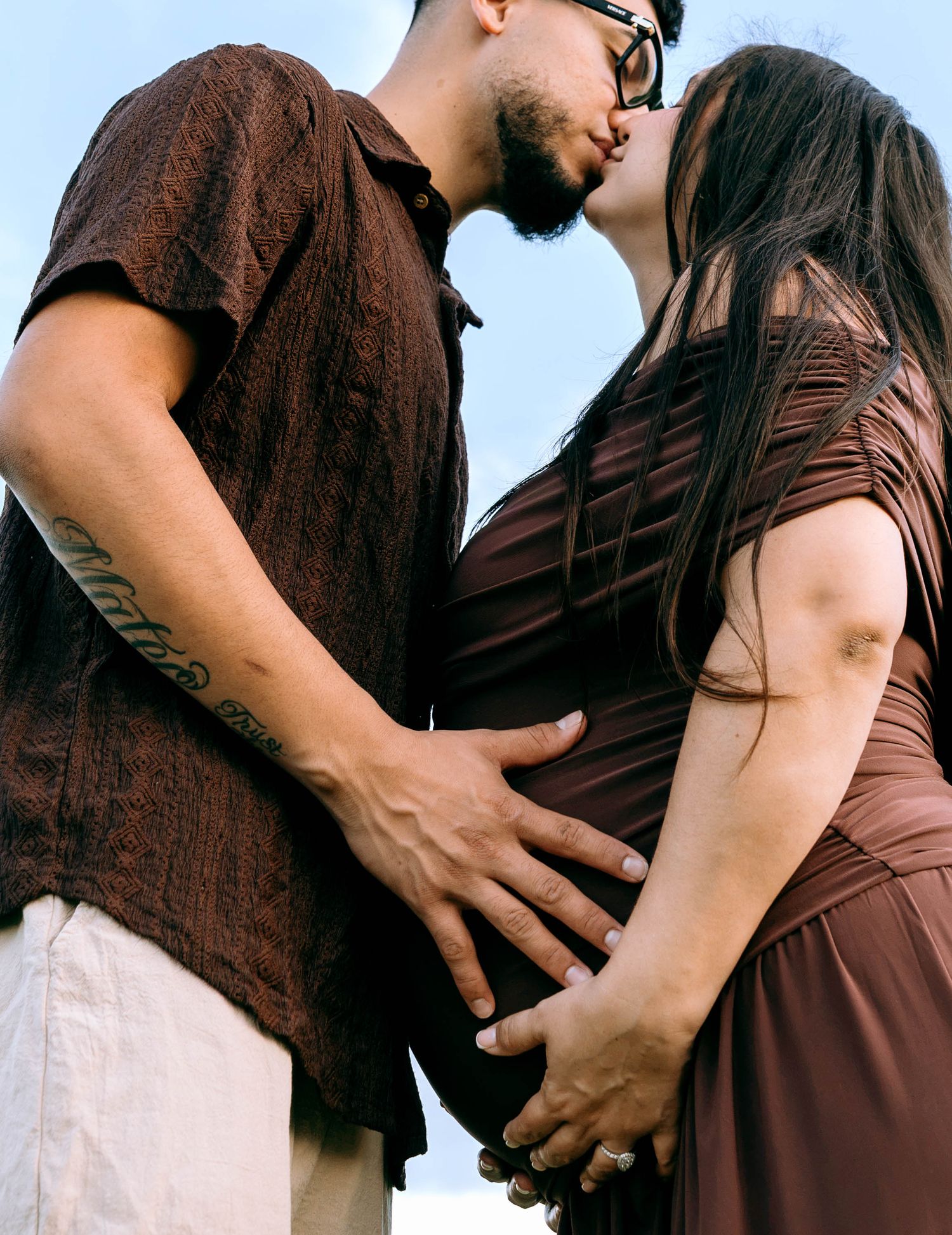 A couple shares a kiss while cradling a pregnant belly outdoors, dressed in matching brown outfits.