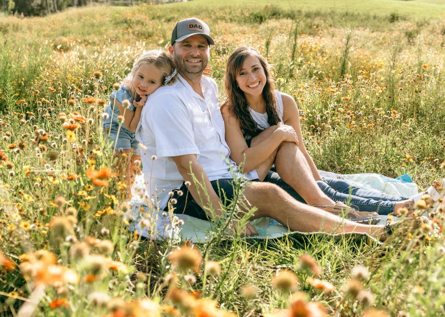 Family of three smiling together in a sunny wildflower meadow, sitting on a blanket surrounded by orange and yellow blooms.