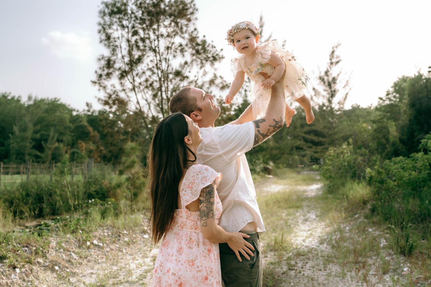 A couple joyfully lifts their baby girl in a sunny outdoor setting surrounded by lush greenery.