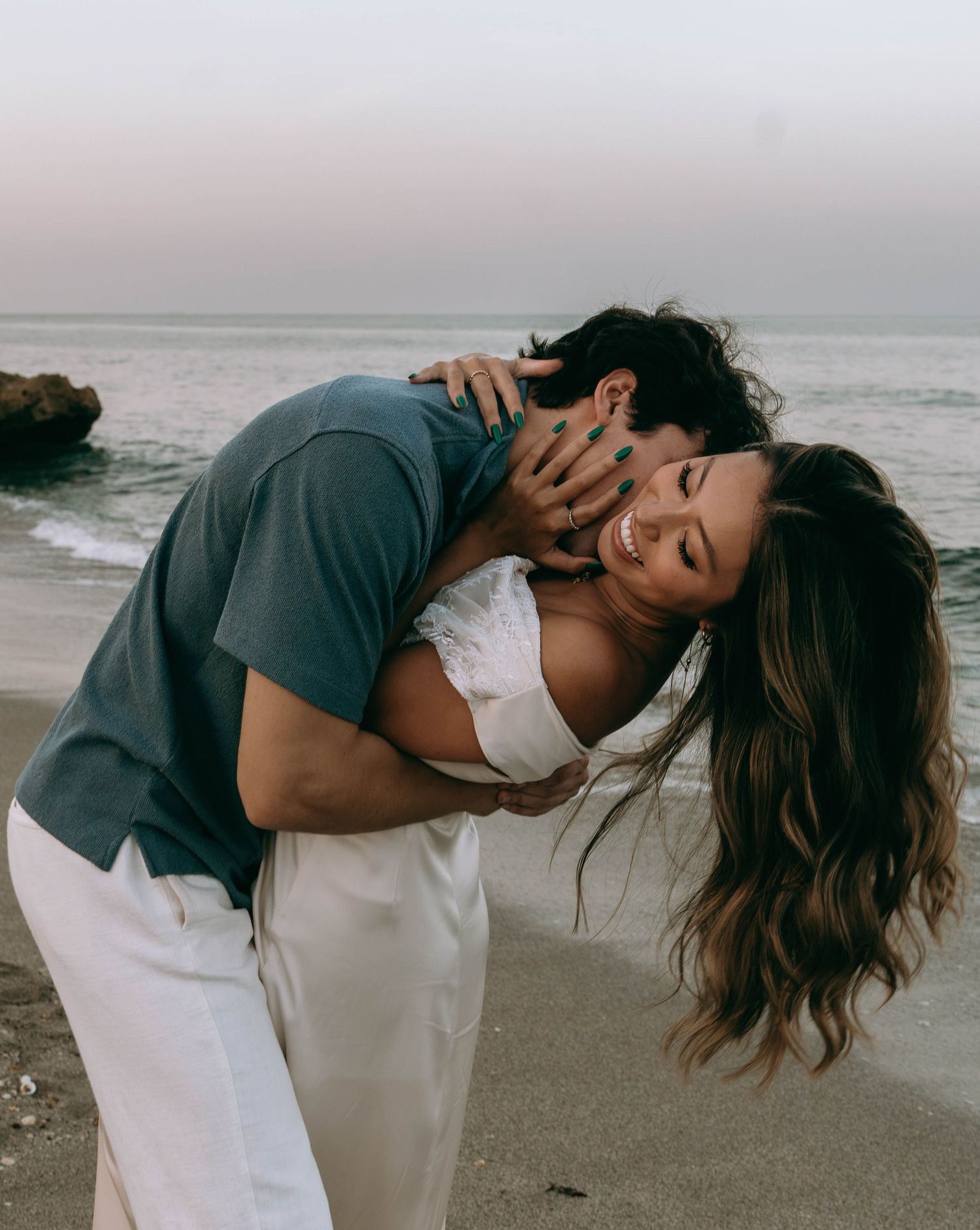 A joyful couple embraces and laughs together on a sandy beach at sunset, with ocean waves in the background.