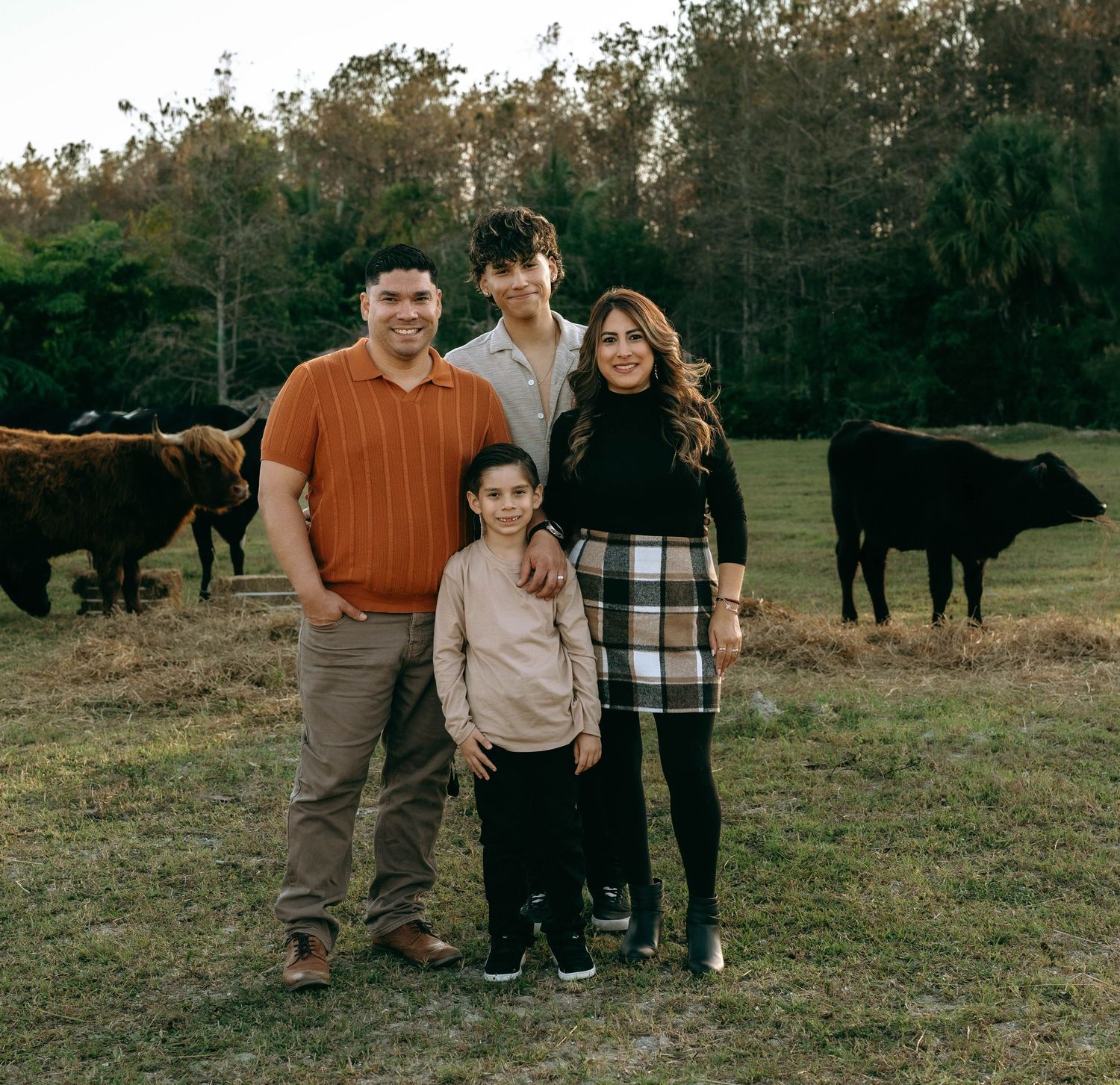 Family of four posing in a field with cattle and trees in the background on a farm.