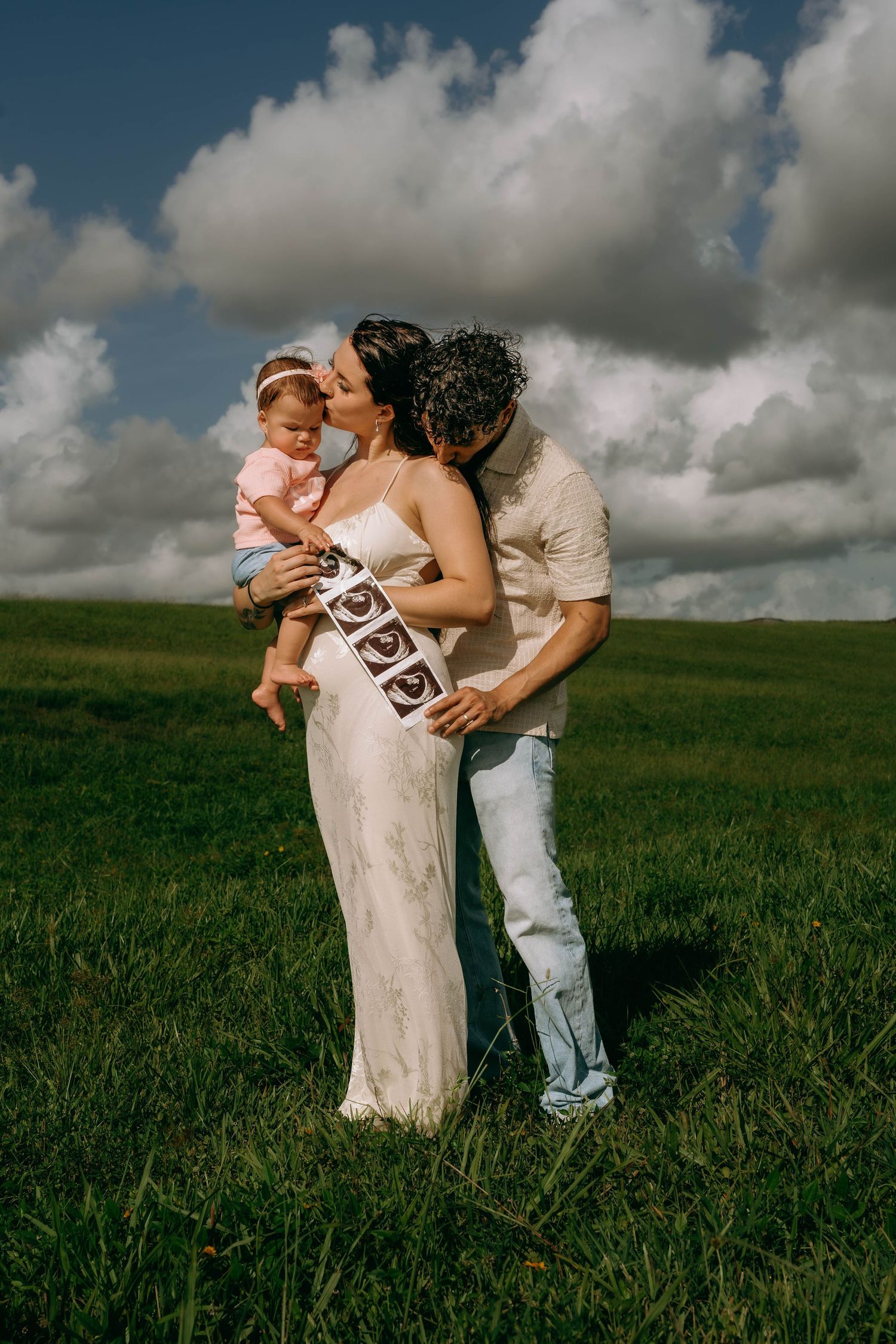 Pregnant couple standing in green field, man kissing woman holding baby, displaying ultrasound photos outdoors.