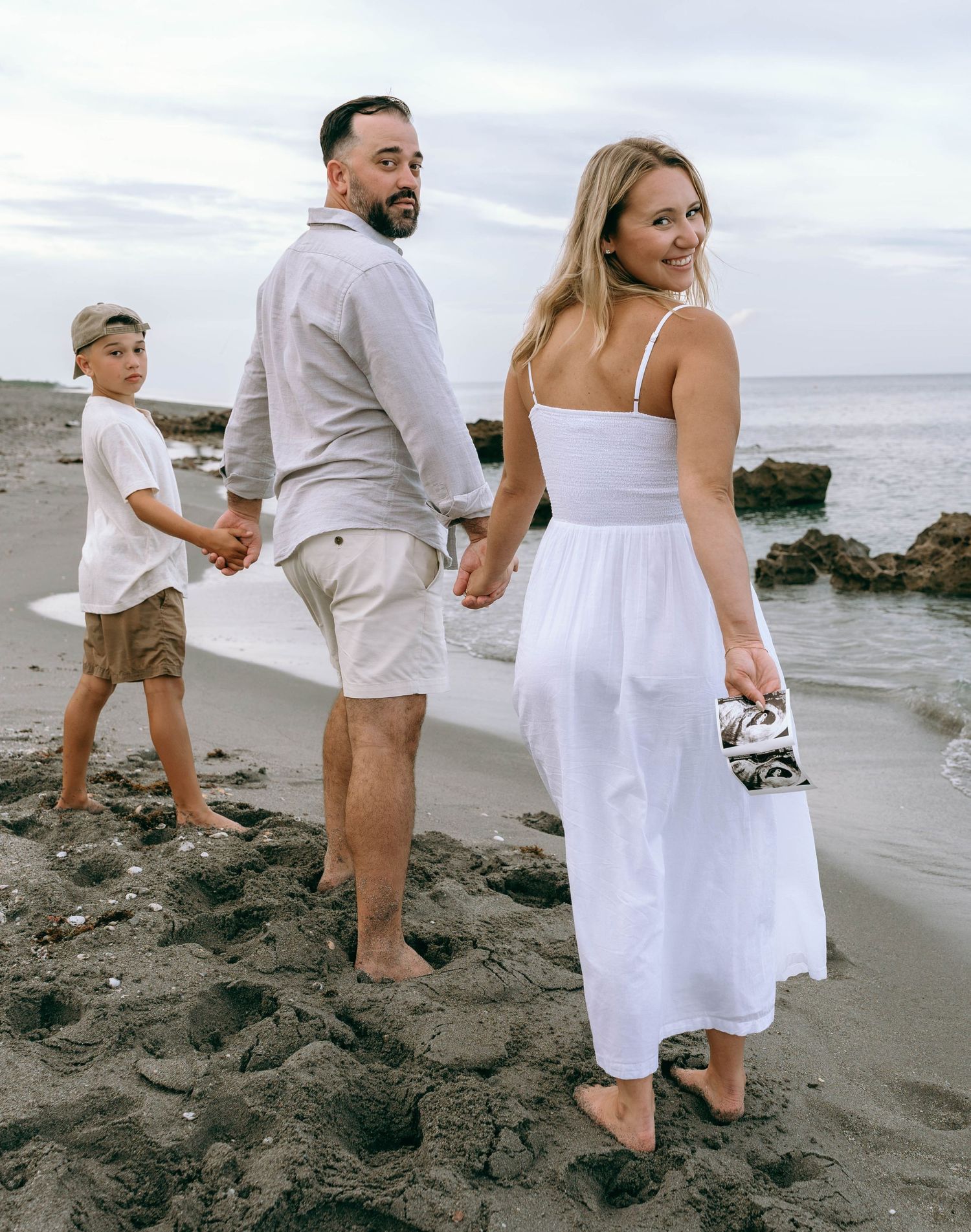 Family of three holding hands on a sandy beach, woman in white dress looking back, man in gray shirt and shorts.