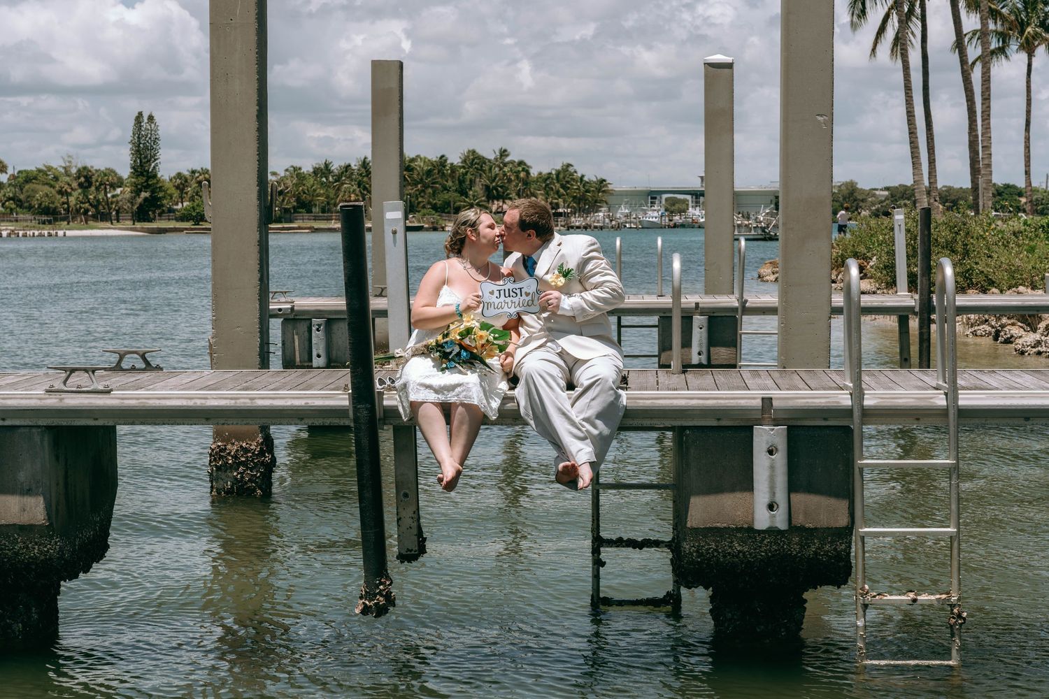 Newlywed couple kissing on a wooden dock over turquoise water with palm trees in the background.