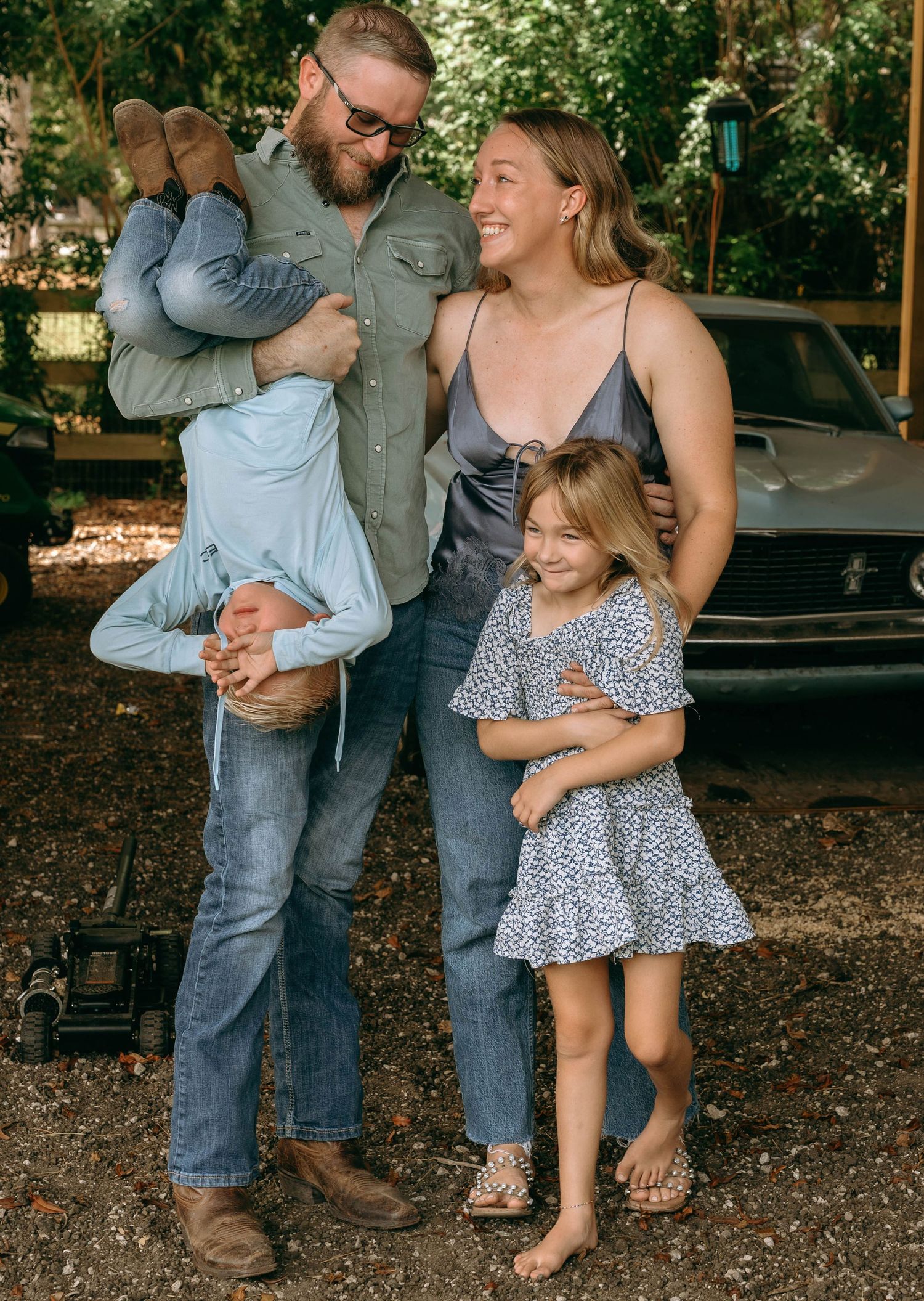 Happy family of four posing outdoors with vintage car, father holding young son upside down while daughter and mother smile.
