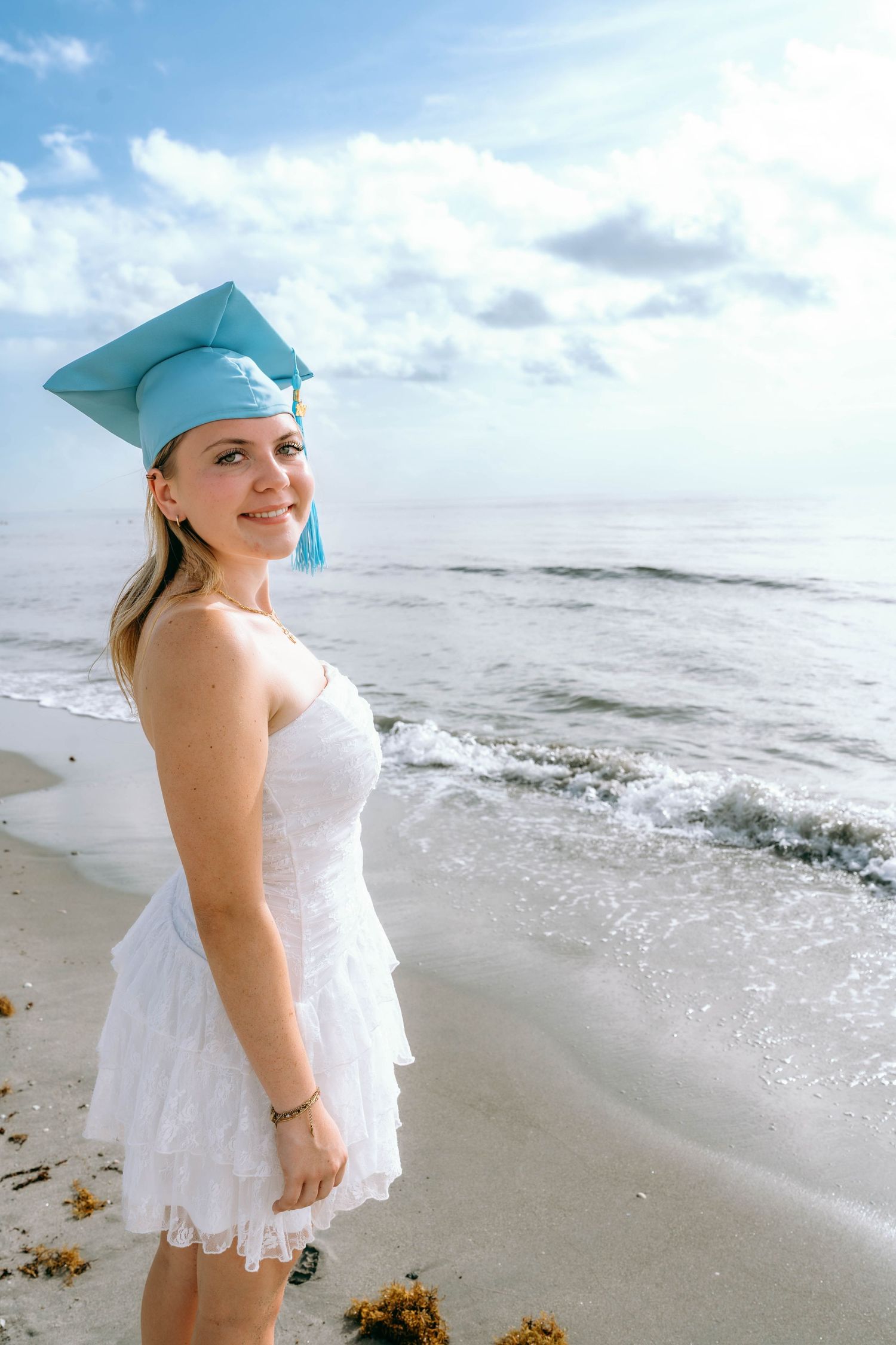 Young woman in white dress and light blue graduation cap smiling on a sunny beach with ocean waves.
