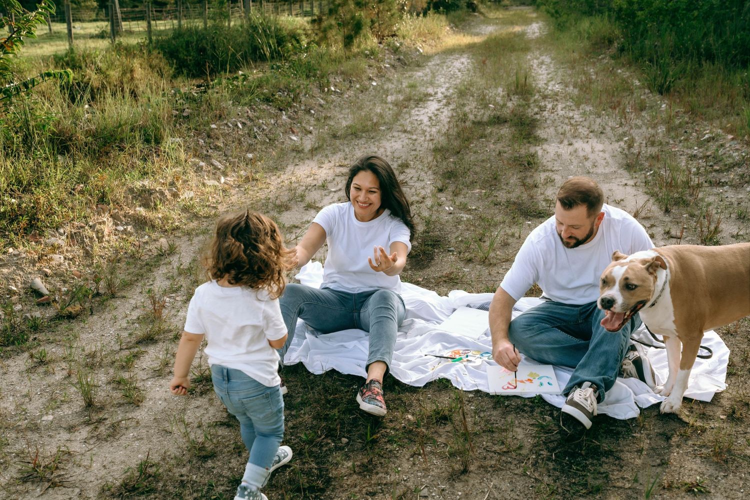 Family with dog enjoying a picnic on a rural dirt path surrounded by greenery on a sunny day.