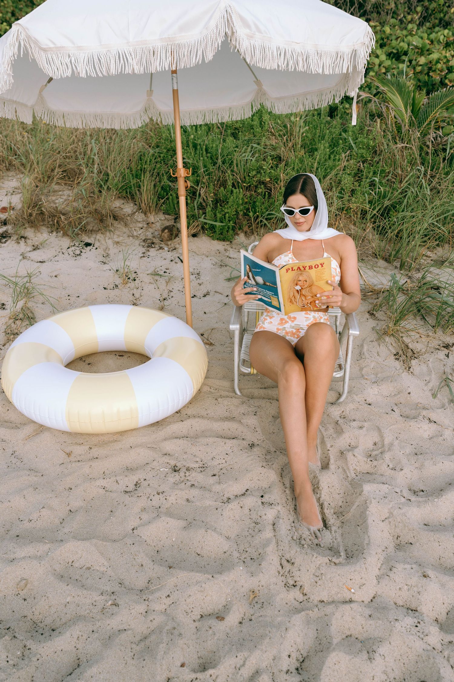 Woman reading a Playboy magazine on a beach chair under a white fringed umbrella beside a yellow striped inflatable ring.