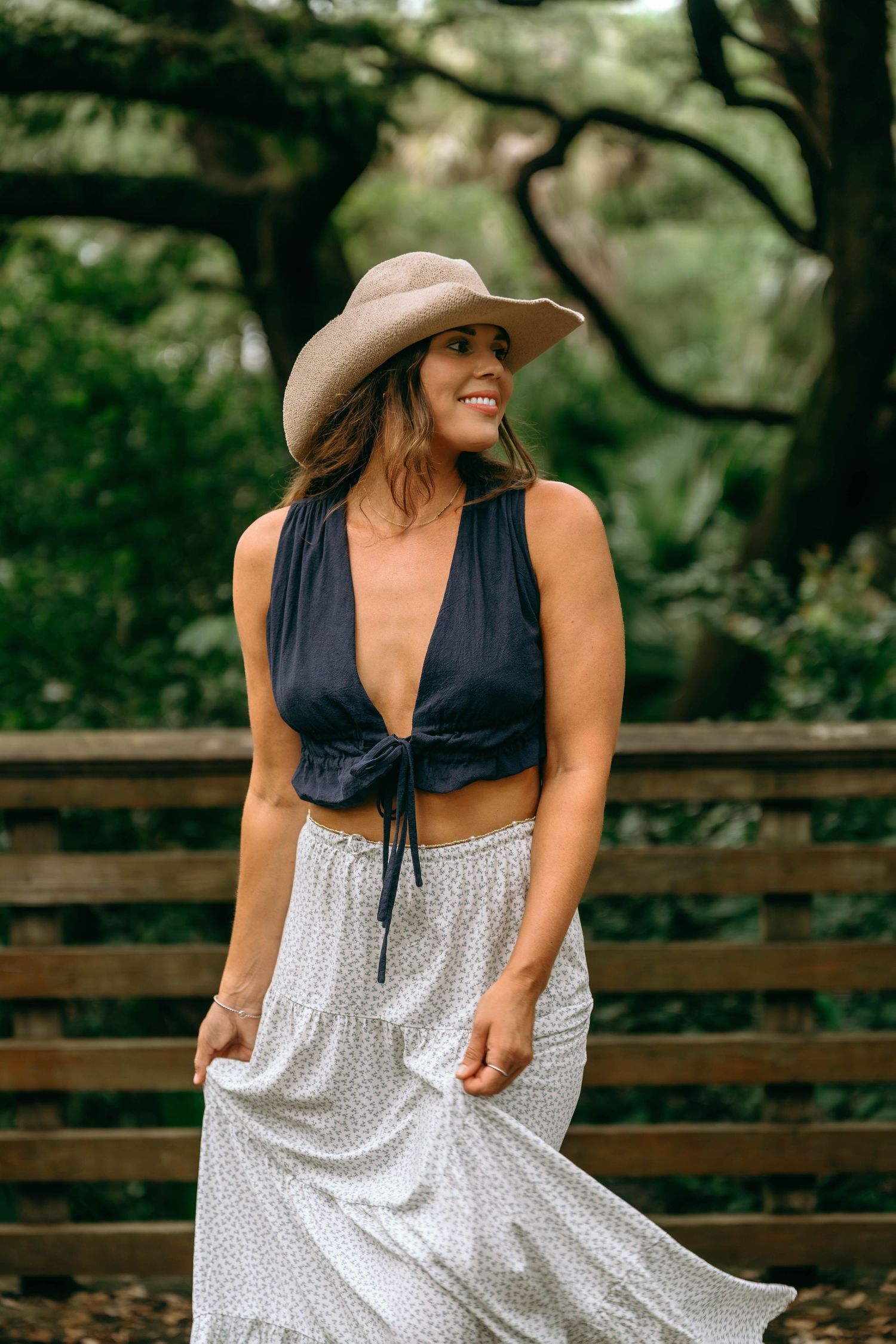 Woman in black crop top, white polka dot skirt, and tan wide-brim hat smiling outdoors near wooden fence.