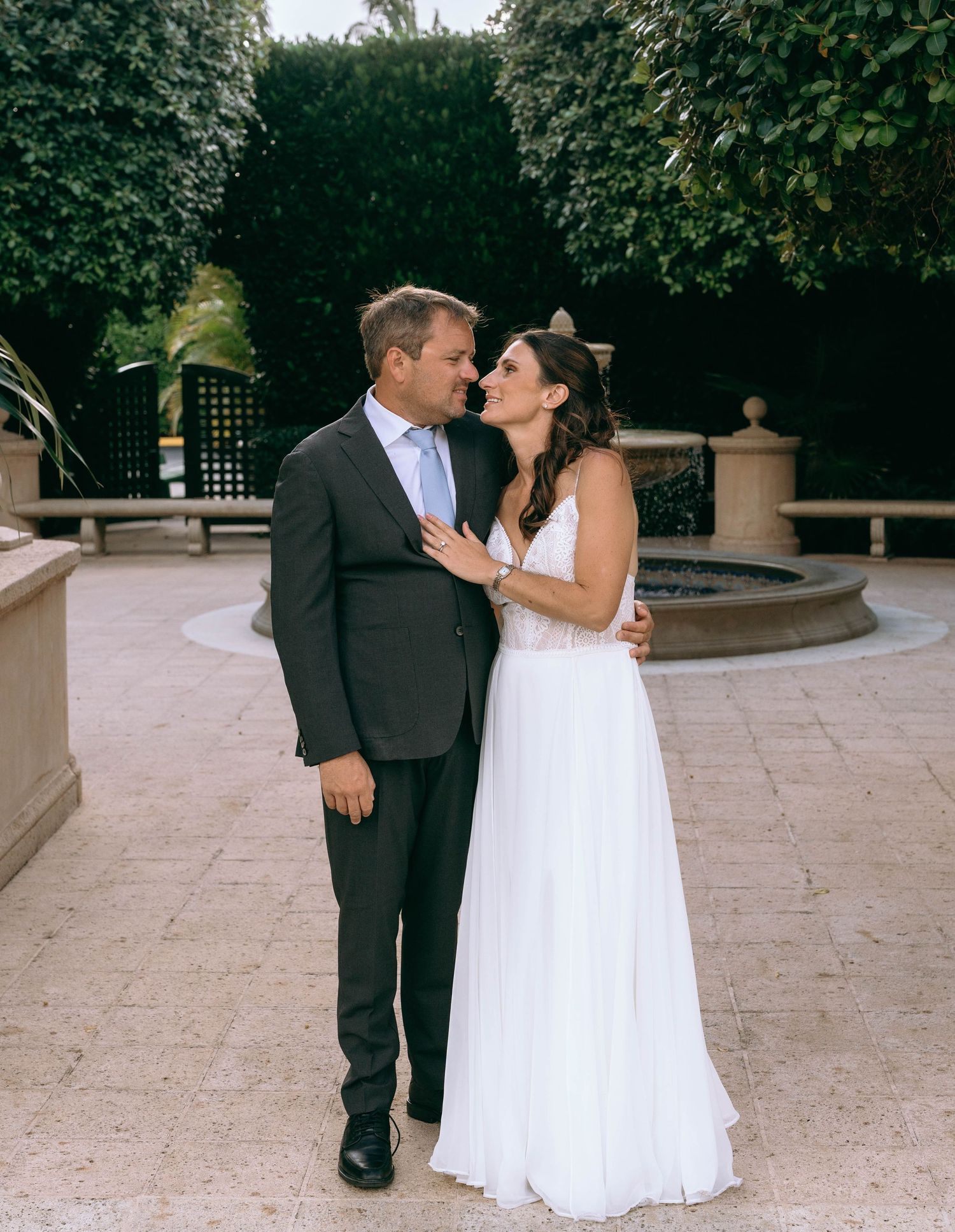 Bride and groom share a romantic moment near a stone fountain in a lush garden wedding venue.
