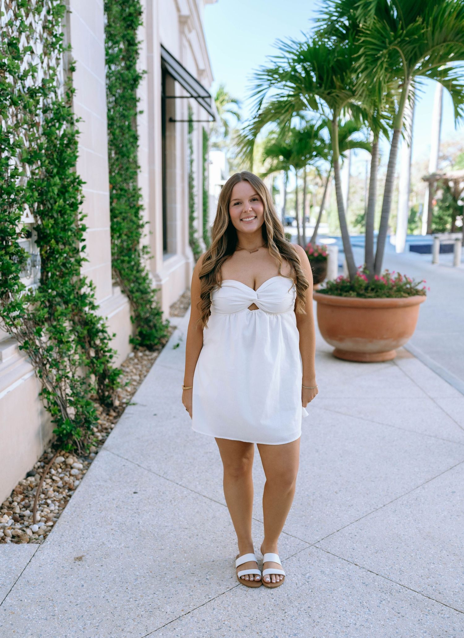 Young woman in white strapless mini dress with cutout detail, smiling on sunny sidewalk with palm trees.