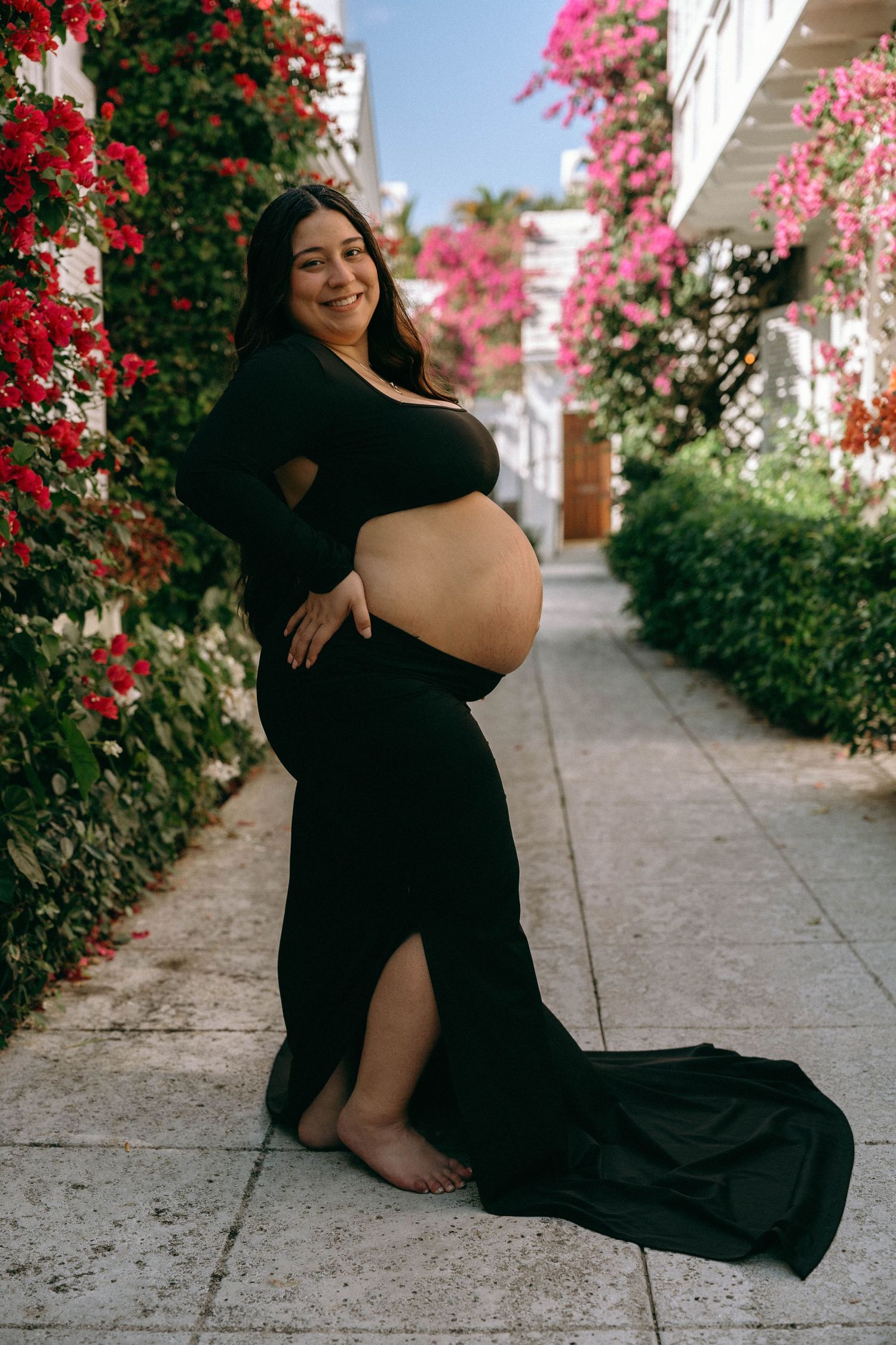 Smiling pregnant woman in elegant black maternity gown posing in vibrant floral garden pathway. Worth Avenue, Palm Beach, FL