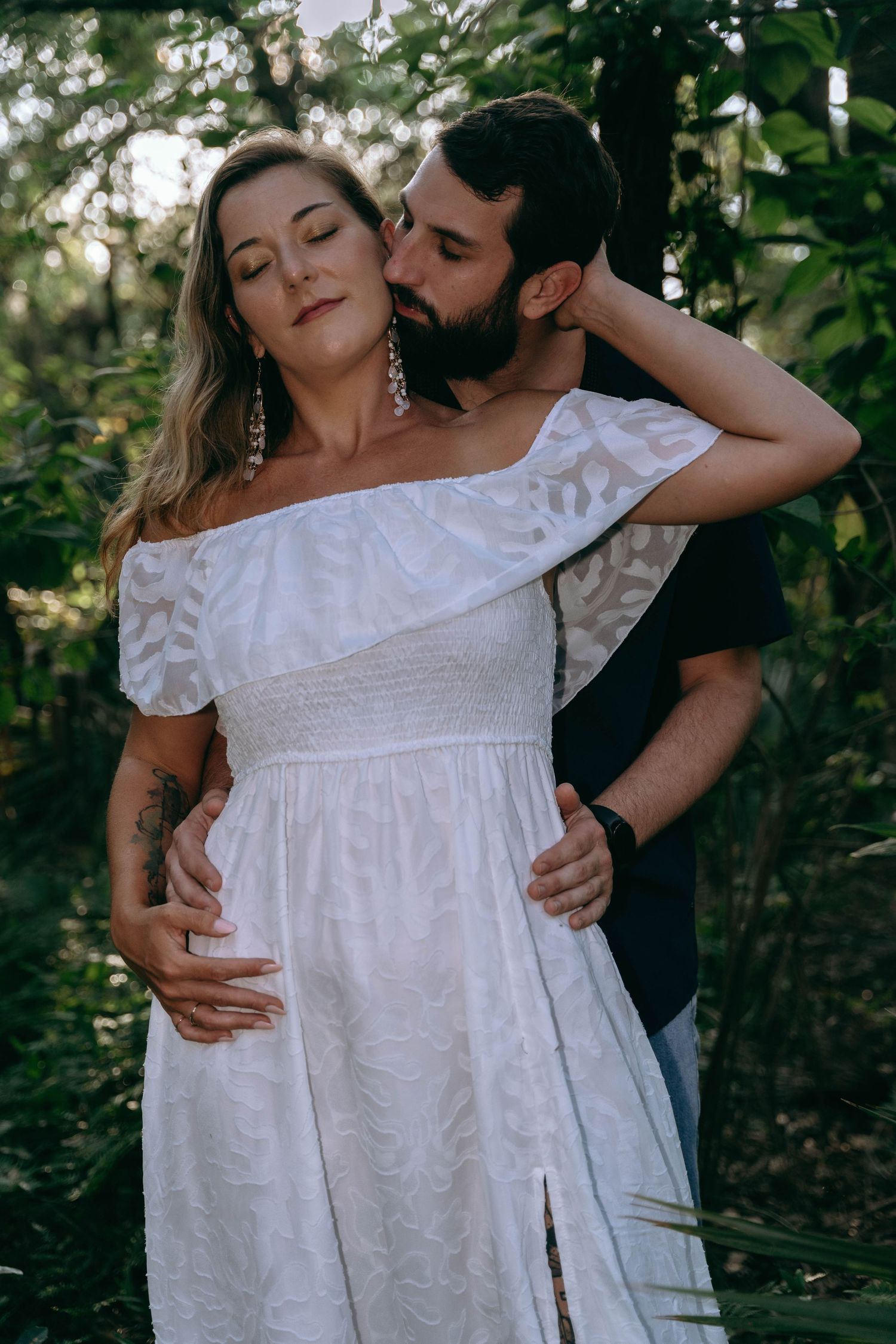 Intimate portrait of couple in white dress and dark clothing against lush green foliage.